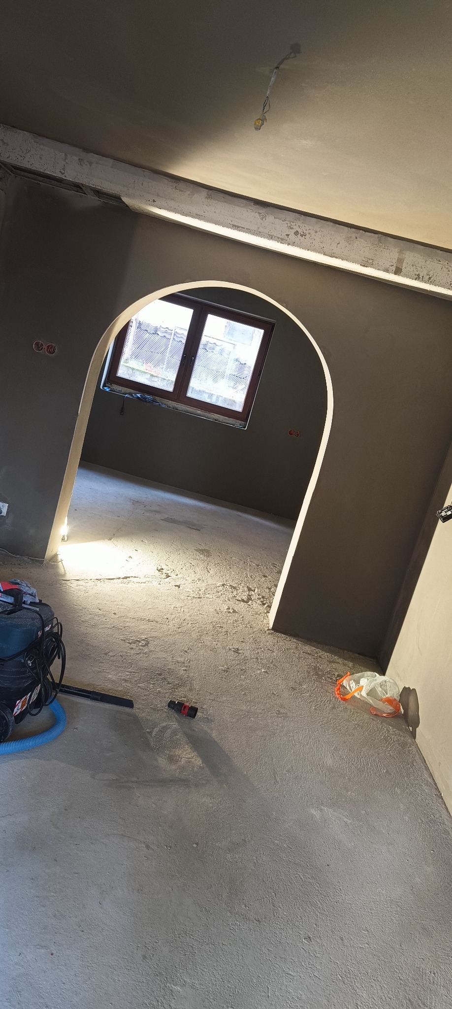 Wnętrze w trakcie prac wykończeniowych: ściany pokryte świeżym, szarym tynkiem, widoczny łuk drzwiowy i okno z widokiem na zewnątrz, na podłodze leży odkurzacz przemysłowy i resztki materiałów...