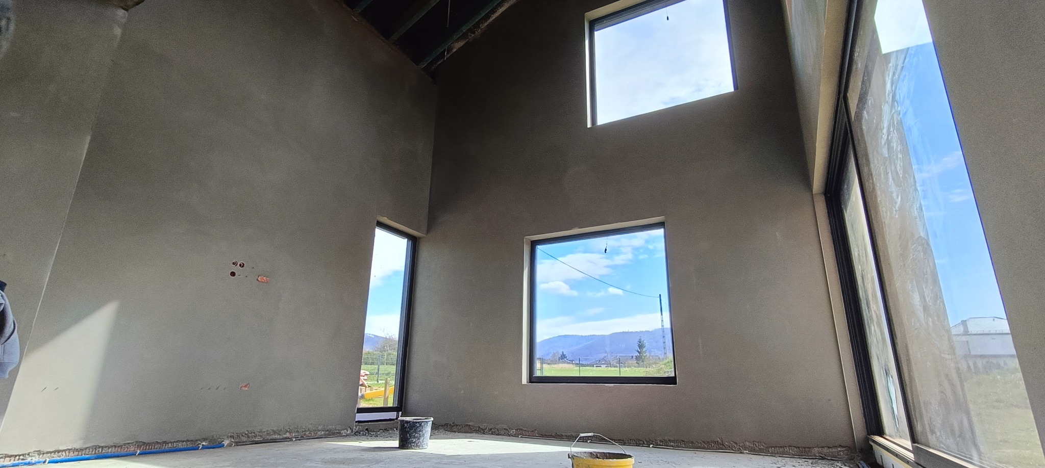 Surowe wnętrze domu w trakcie prac wykończeniowych, ściany pokryte świeżym, szarym tynkiem, widoczne okna z widokiem na krajobraz, dwa wiadra na podłodze, niebieskie rurki przy podłodze.