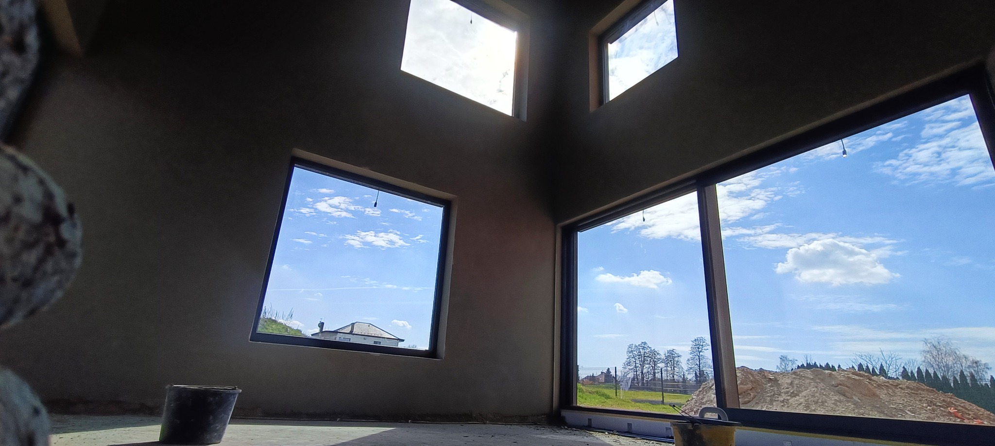 Wnętrze domu w stanie surowym po nałożeniu tynków maszynowych, widoczne okna z widokiem na błękitne niebo i okolicę, kubeł na podłodze.