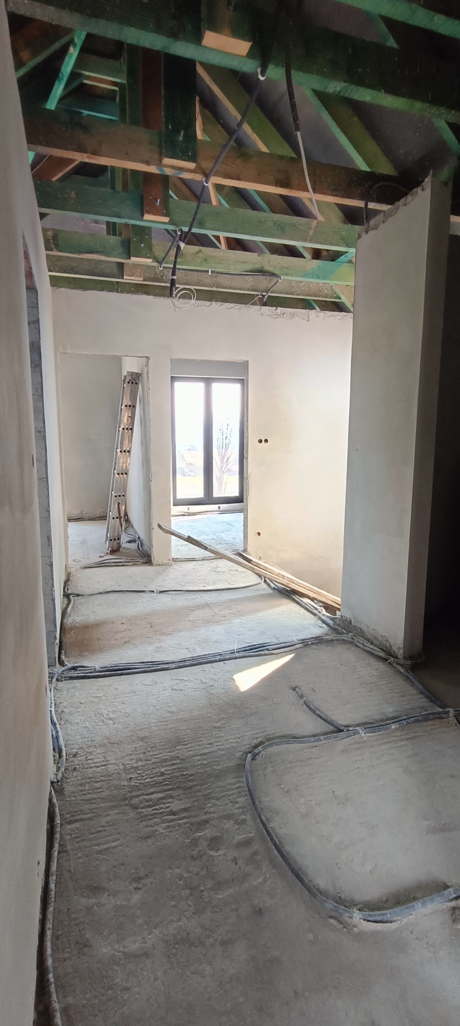 Wnętrze domu w trakcie prac wykończeniowych: surowe ściany przygotowane do dalszych prac, widoczne instalacje elektryczne na suficie i ogrzewanie podłogowe, drabina oparta o ścianę w oddali.