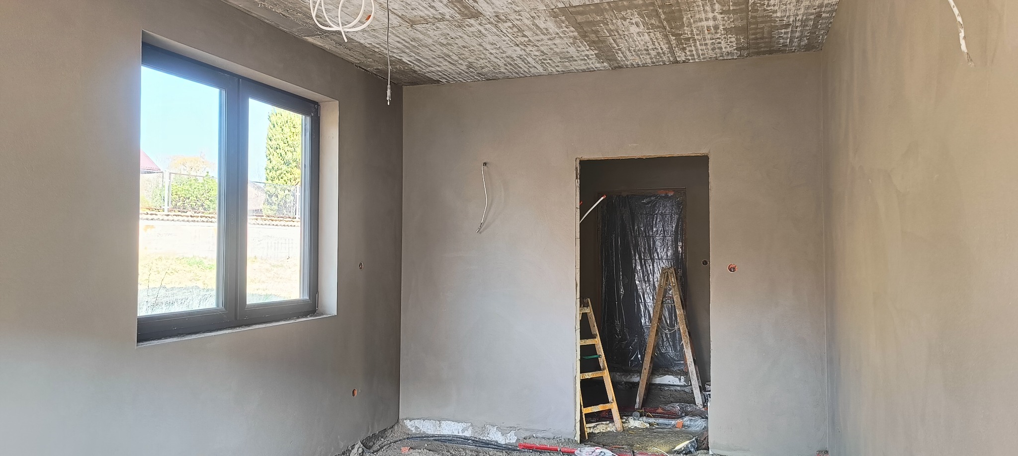 Wnętrze pokoju po nałożeniu szarego tynku, widoczne okno z widokiem na zewnątrz, przewody elektryczne na suficie oraz drabina oparta o wejście do innego pomieszczenia zabezpieczonego folią.