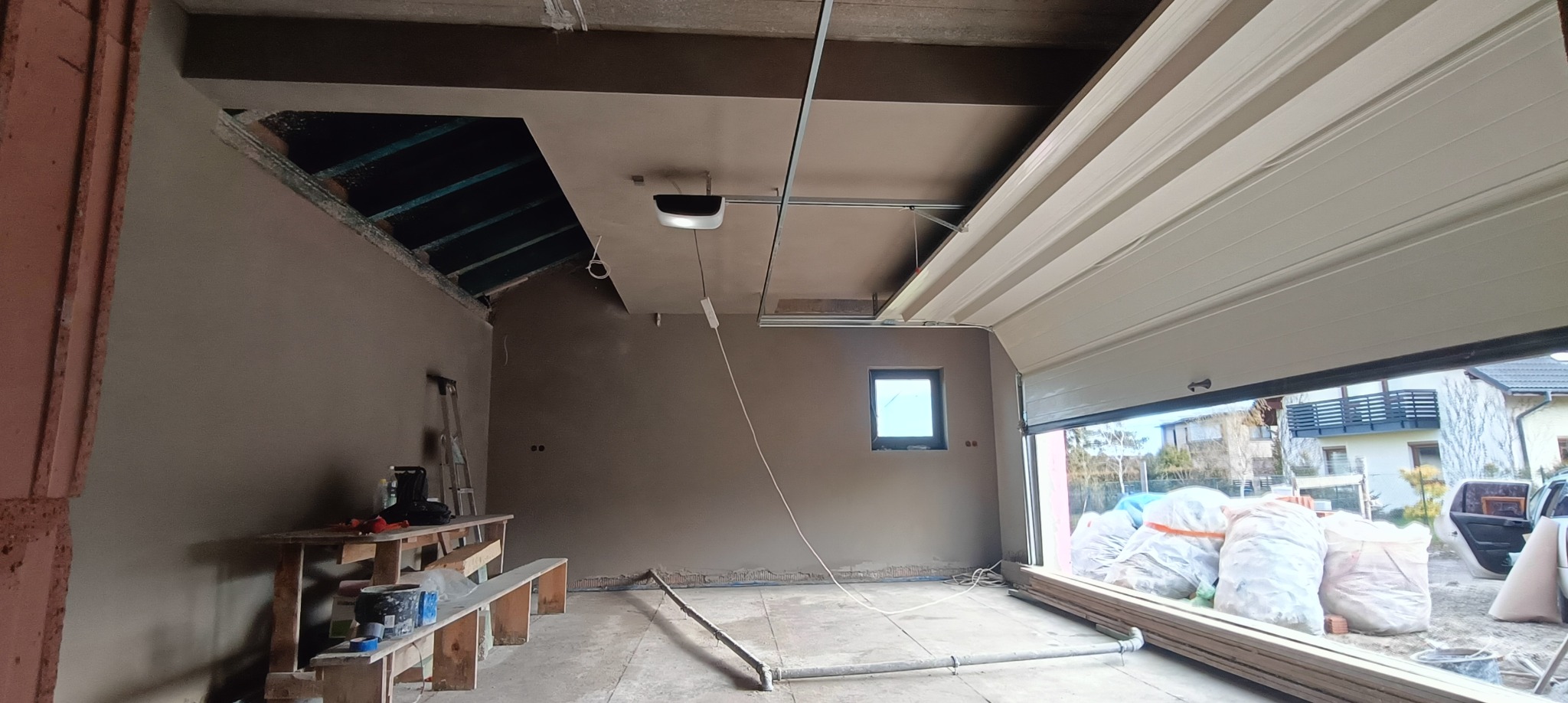 Wnętrze garażu w trakcie prac wykończeniowych: ściany i sufit pokryte świeżym, szarym tynkiem, widoczne niedociągnięcia przy podłodze, fragment odsłoniętej konstrukcji dachu, otwarta brama garażowa...