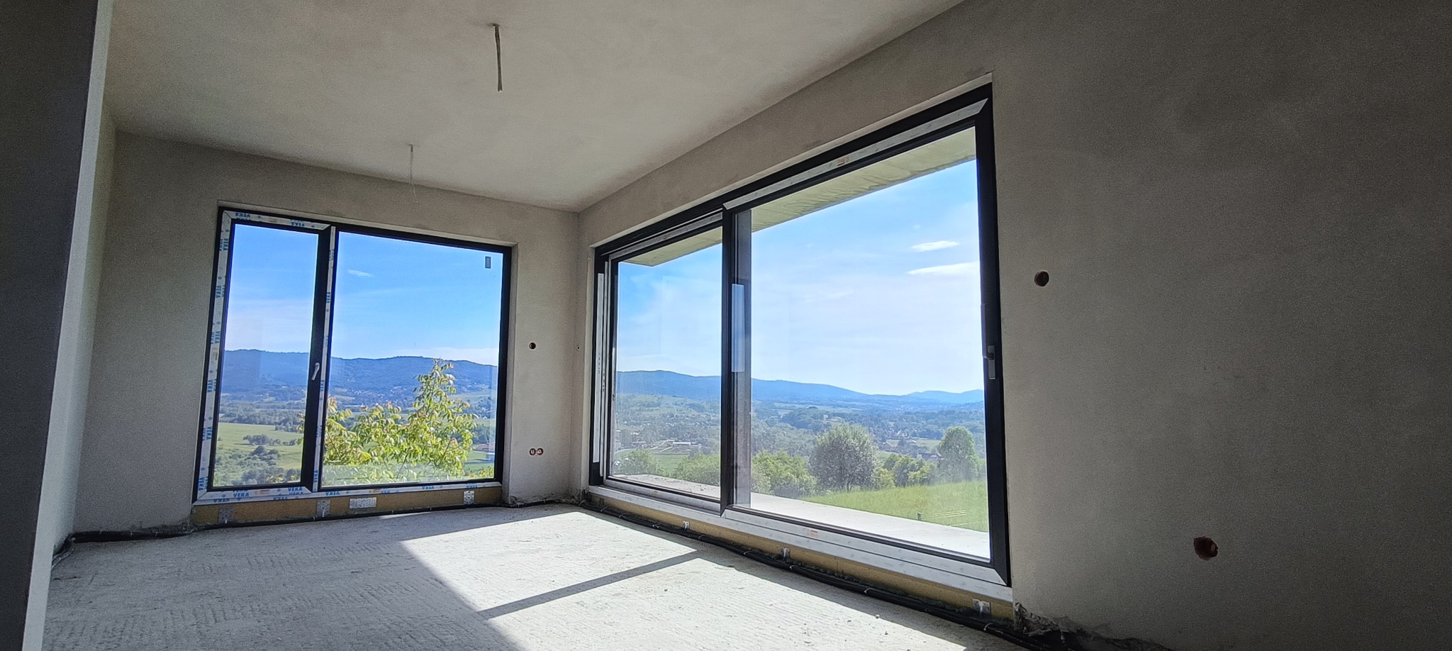 Surowe wnętrze z dwoma dużymi oknami przesuwnymi, przez które widać malowniczy krajobraz wzgórz i lasów. Ściany przygotowane do wykończenia, podłoga surowa.