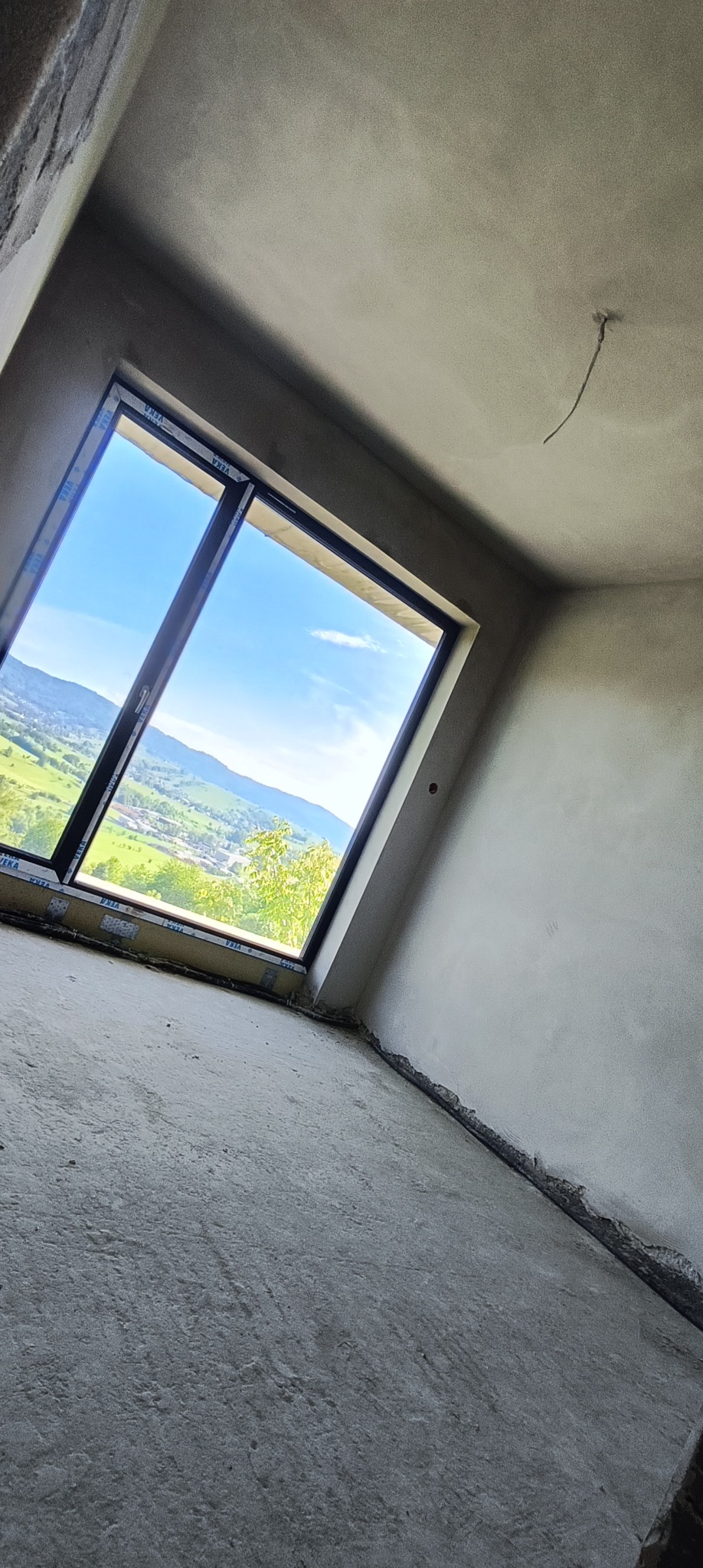 Pokój po tynkowaniu maszynowym, z widokiem na góry przez okno. Świeże tynki na ścianach i suficie, surowy beton na podłodze. Widoczne detale okna.