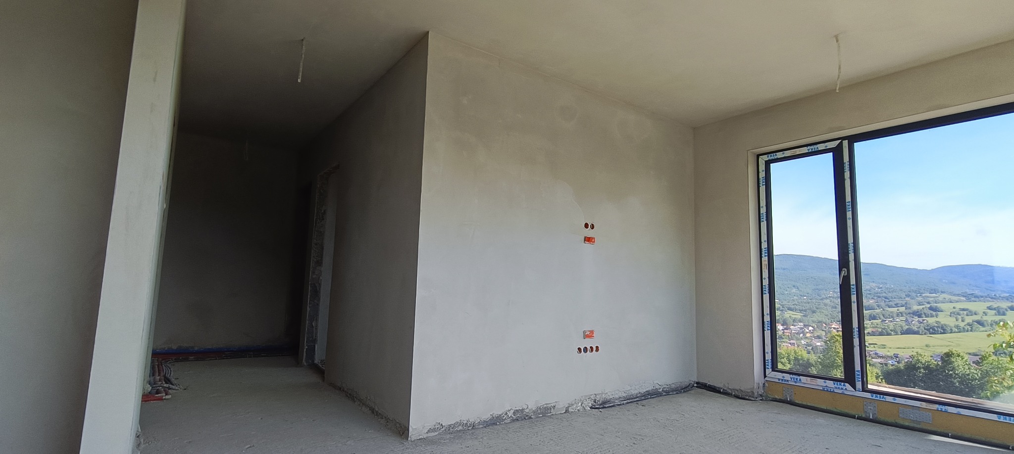 Wnętrze po szpachlowaniu z widocznymi puszkami elektrycznymi, gładzią na ścianach i oknem z widokiem na góry. Podłoga surowa, wylewka.