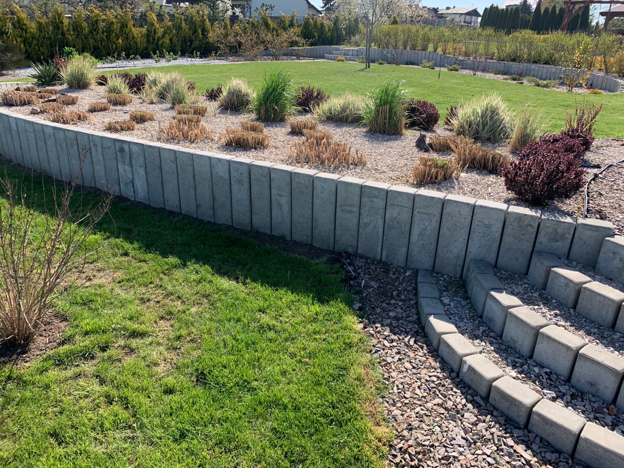 Uporządkowany ogród z rabatami oddzielonymi betonowymi obrzeżami, trawnik i kamienne schody prowadzące w górę.