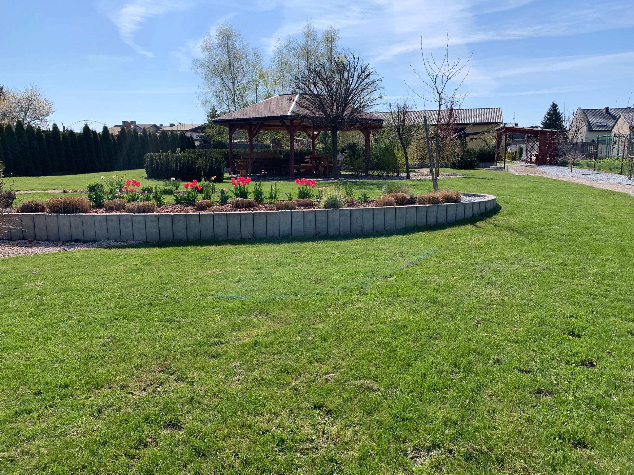 Zadbany trawnik z rabatą kwiatową w betonowej obudowie i altaną ogrodową w tle, widok na posesję w słoneczny dzień.