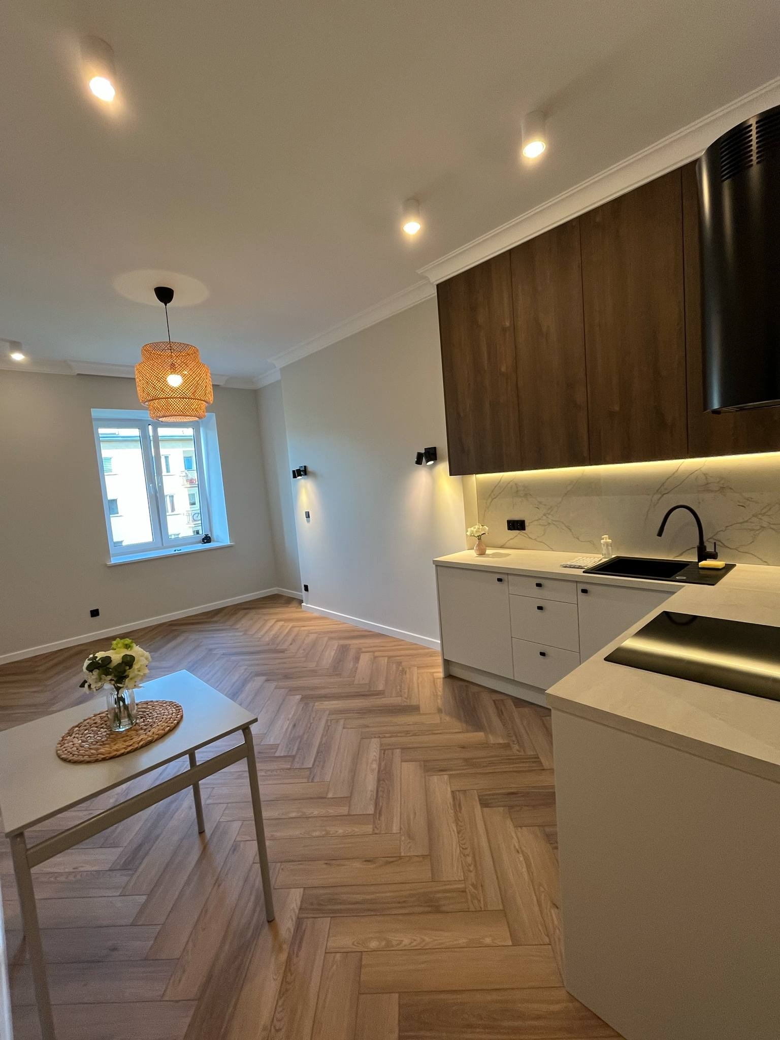 Nowoczesna kuchnia z drewnianą podłogą w jodełkę, białymi szafkami i marmurowym blatem oświetlonym ledami. W tle okno z widokiem na miasto i lampa z plecionki.