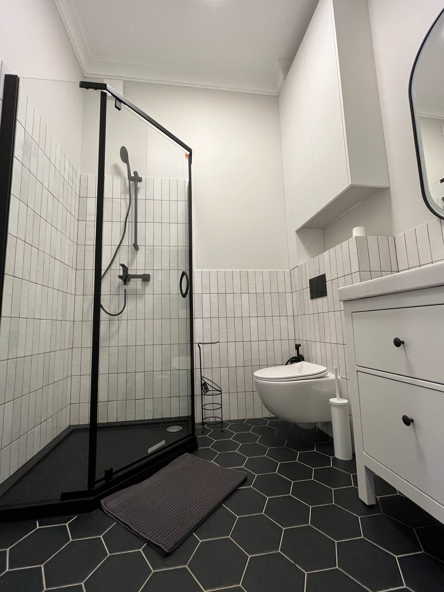 Nowoczesna łazienka z czarną kabiną prysznicową typu walk-in, heksagonalną podłogą i białymi płytkami ułożonymi pionowo. Minimalistyczny design z czarnymi akcentami.