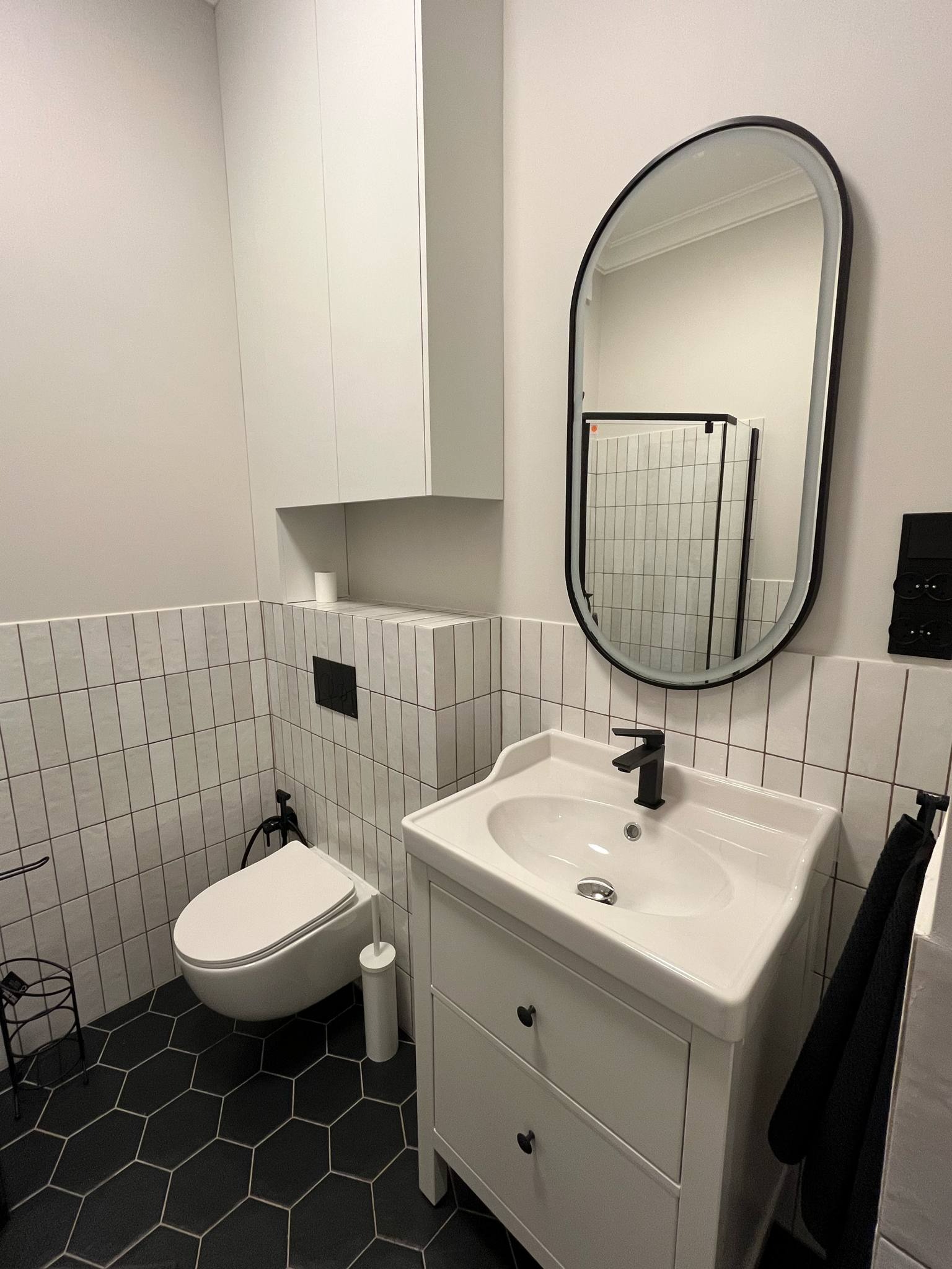 Nowoczesna łazienka z białymi płytkami, czarną armaturą i heksagonalną podłogą. Umywalka z szafką, owalne lustro w czarnej ramie i wisząca szafka nad toaletą.