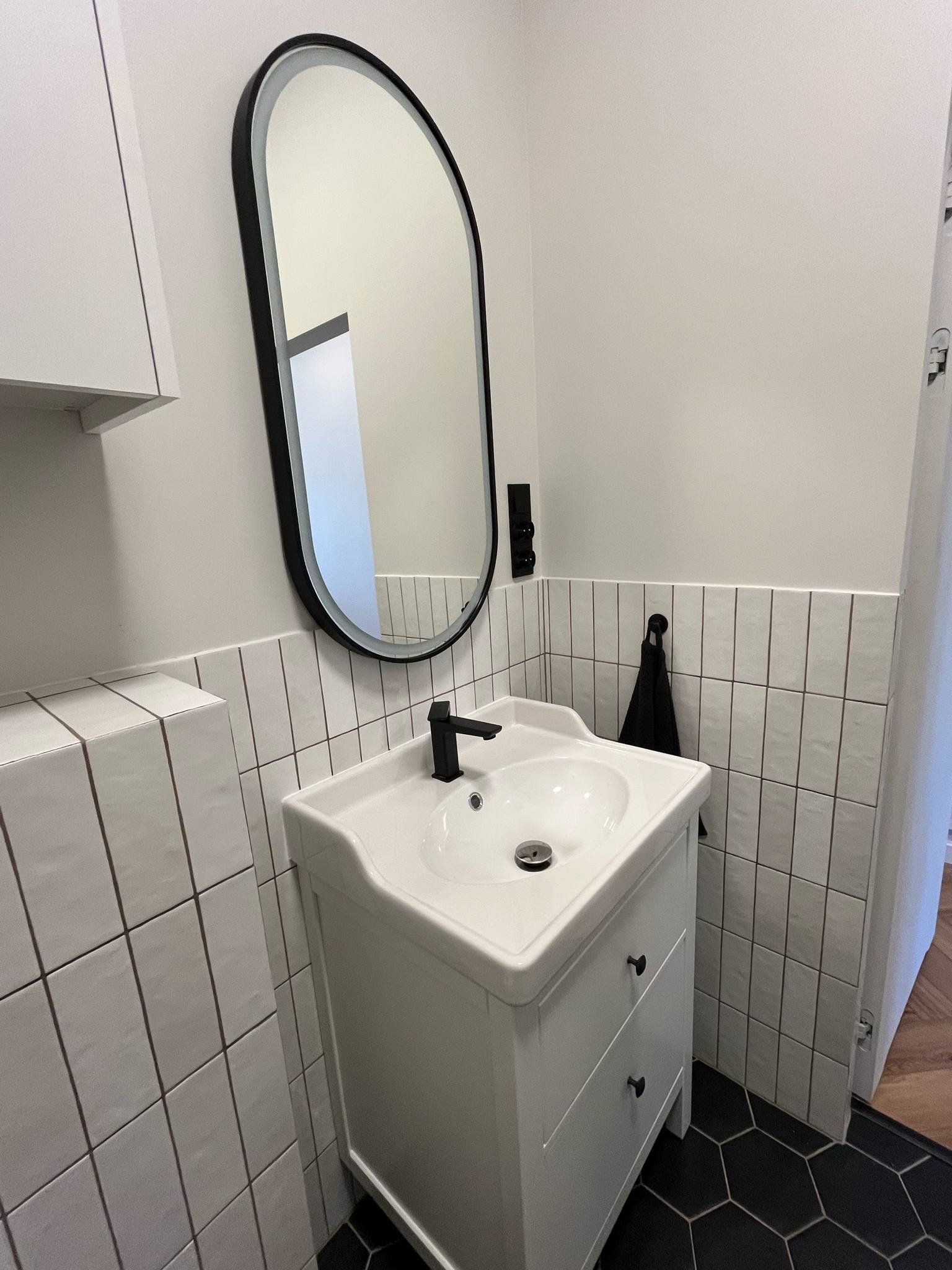 Biała umywalka z czarną armaturą i owalnym lustrem w nowoczesnej łazience z czarną, heksagonalną podłogą i białymi płytkami imitującymi cegłę.