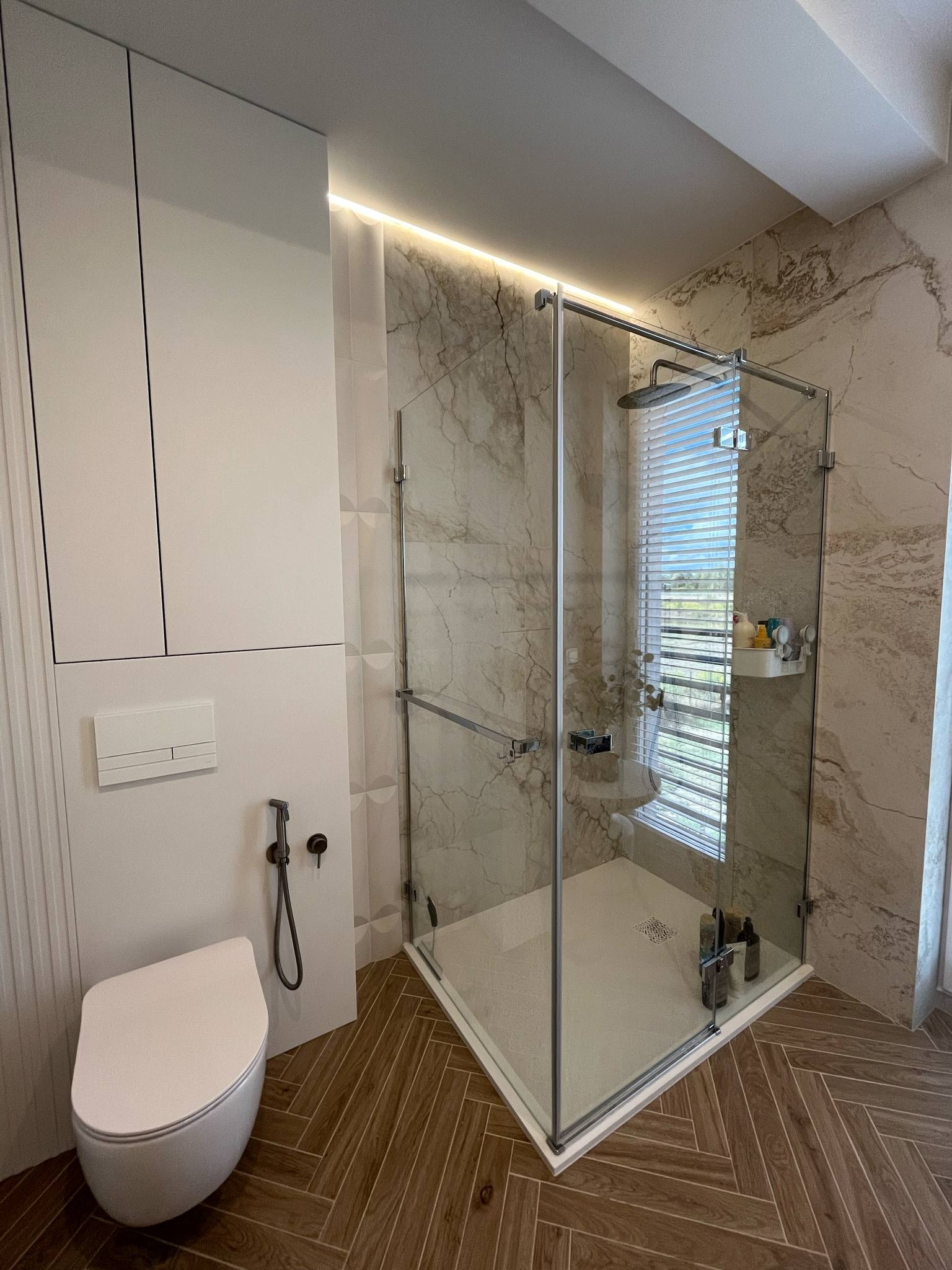 Nowoczesna łazienka z kabiną prysznicową typu walk-in, białym WC podwieszanym i drewnopodobną podłogą ułożoną w jodełkę. Marmurowe płytki na ścianach i subtelne oświetlenie LED.