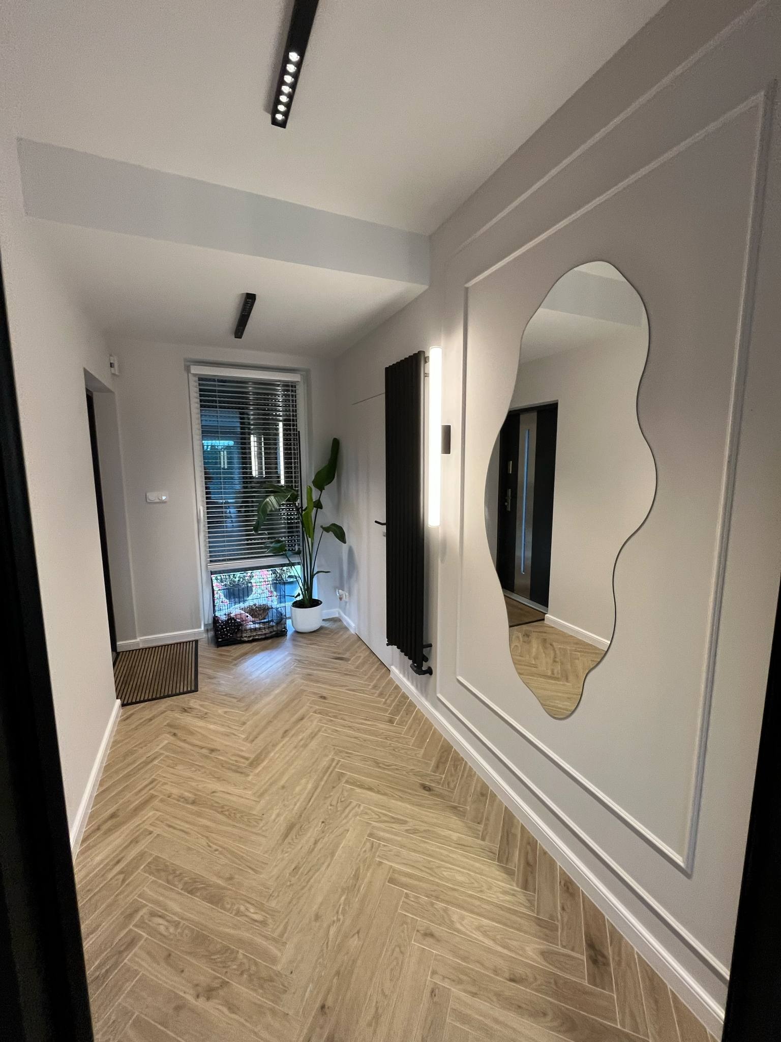 Stylowy przedpokój z parkietem w jodełkę, asymetrycznym lustrem w ozdobnej ramie, grzejnikiem i oświetleniem LED. Widok na okno z żaluzjami i rośliną.