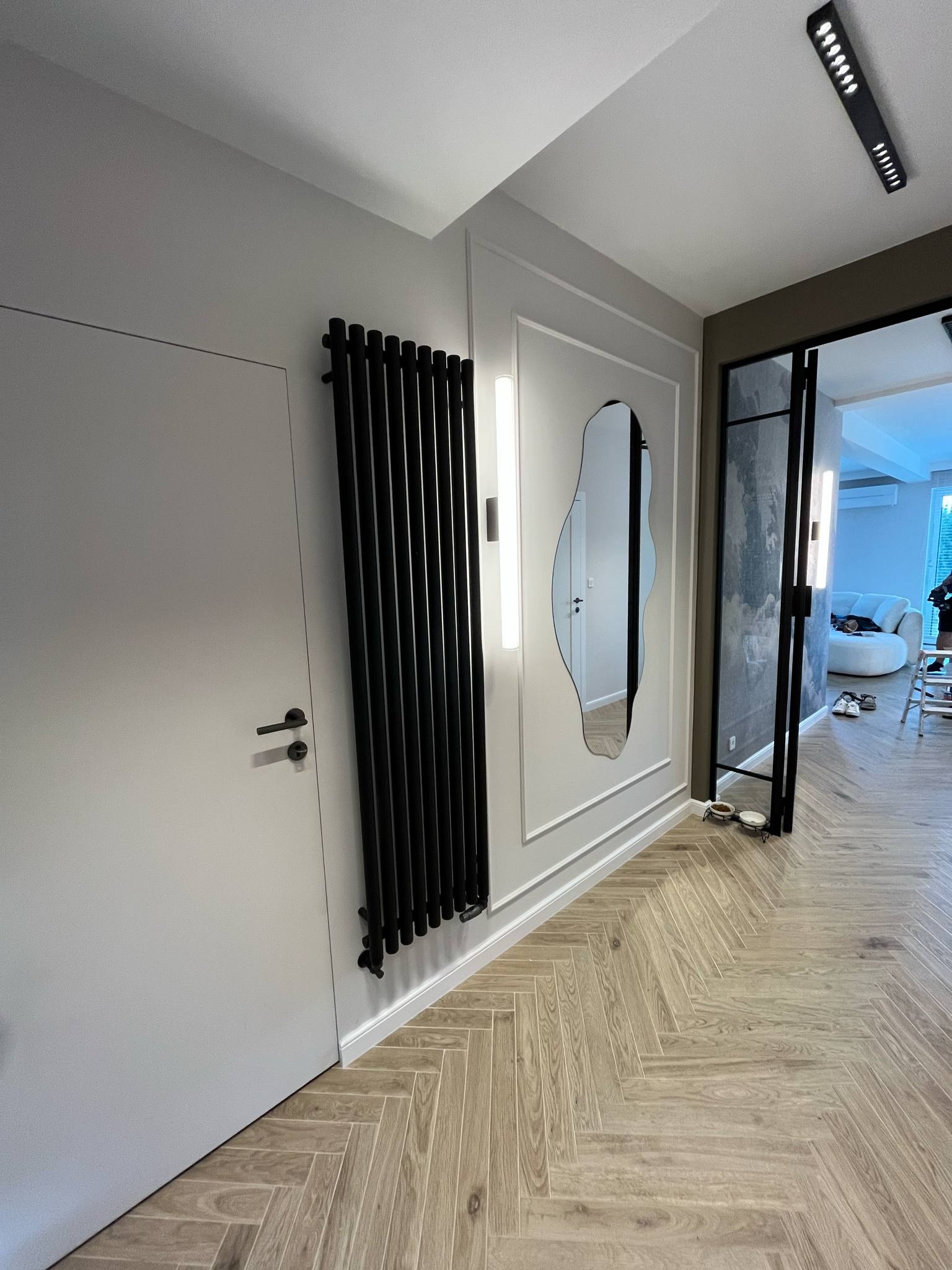 Nowoczesny przedpokój: szara ściana z ukrytym wejściem, czarny grzejnik panelowy, asymetryczne lustro w ozdobnej ramie i parkiet ułożony w jodełkę.