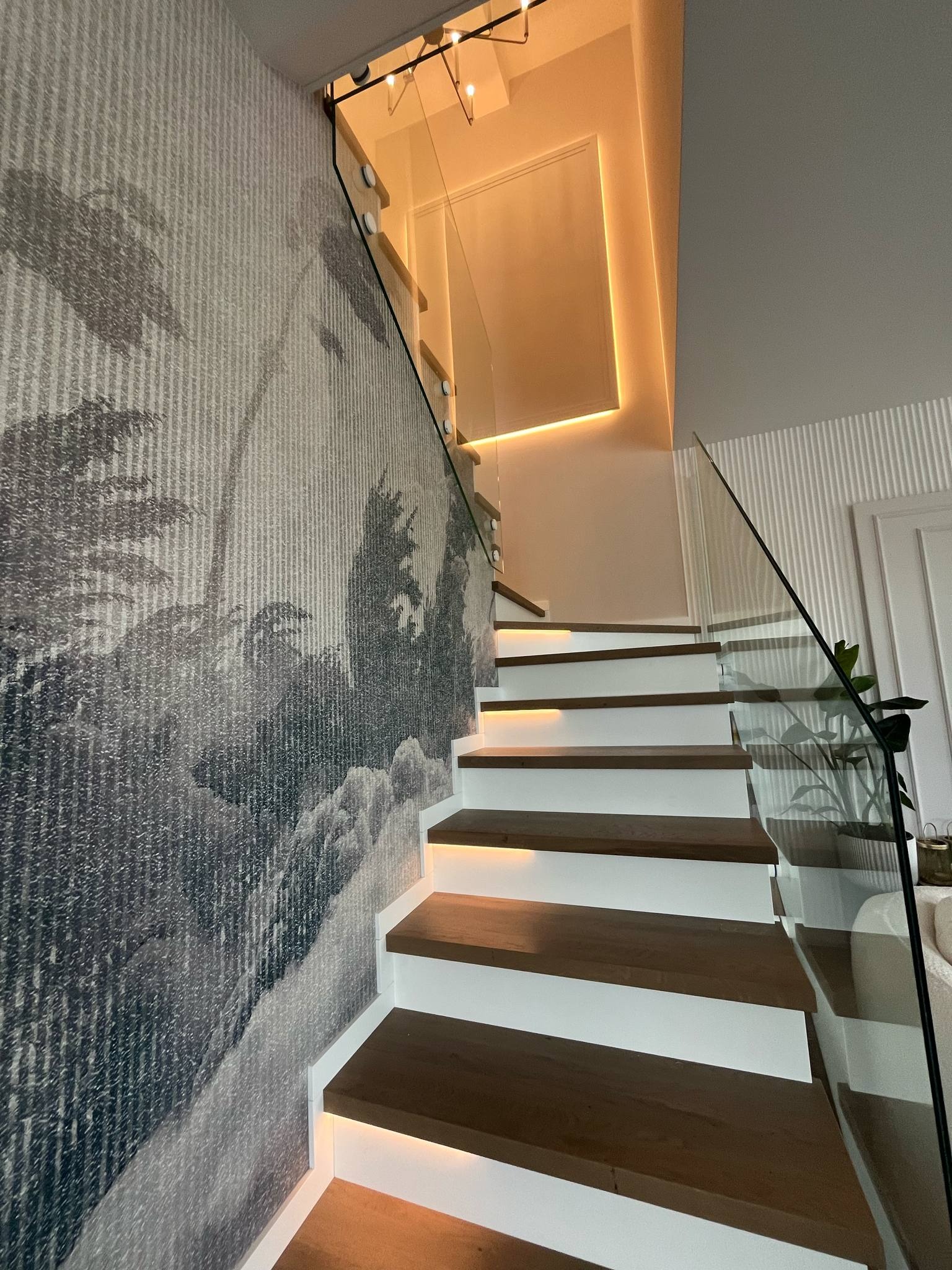 Nowoczesne schody z drewnianymi stopniami i białymi podstopnicami, podświetlane. Szklana balustrada i tapeta w tropikalny wzór. Oświetlenie ledowe na suficie.