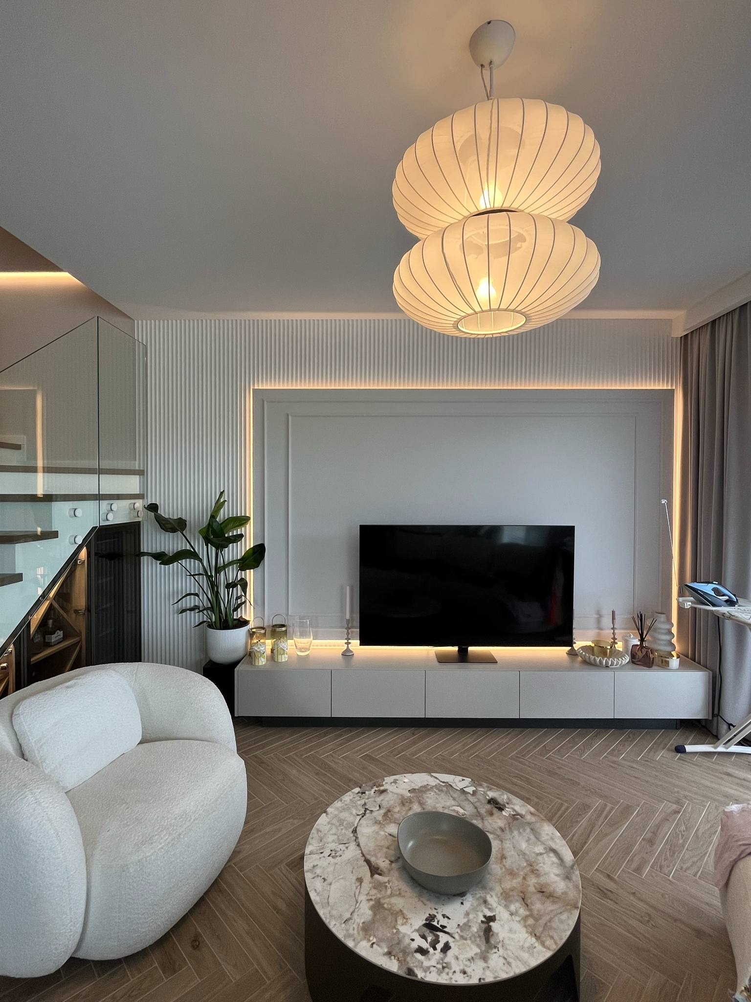 Jasne wnętrze salonu z parkietem w jodełkę, białym fotelem typu baranek, marmurowym stolikiem kawowym i oświetleniem LED. Schody ze szklaną balustradą.