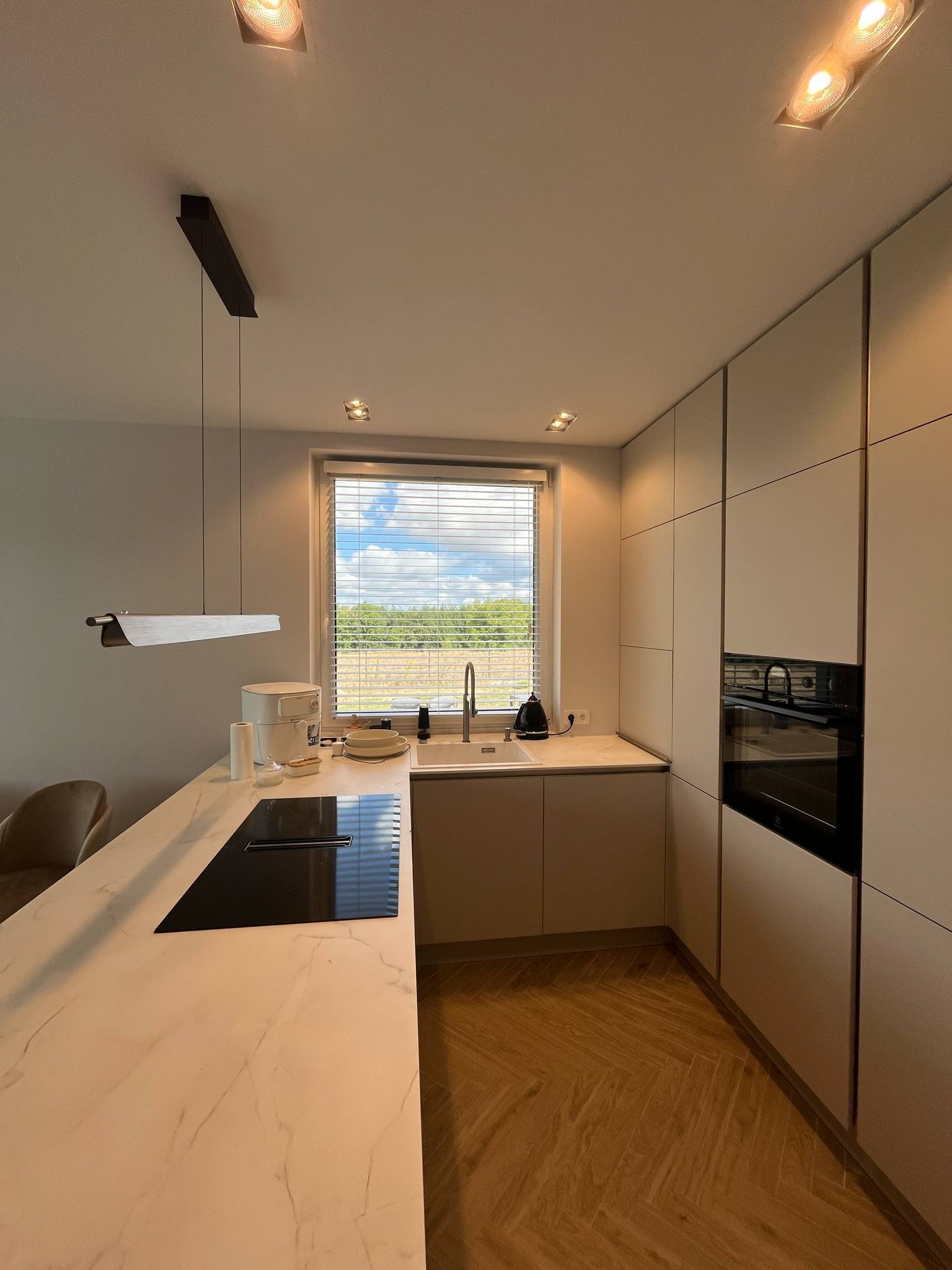 Nowoczesna kuchnia z marmurowym blatem, płytą indukcyjną i widokiem na zieleń za oknem. Minimalistyczne szafki i drewniana podłoga w jodełkę.