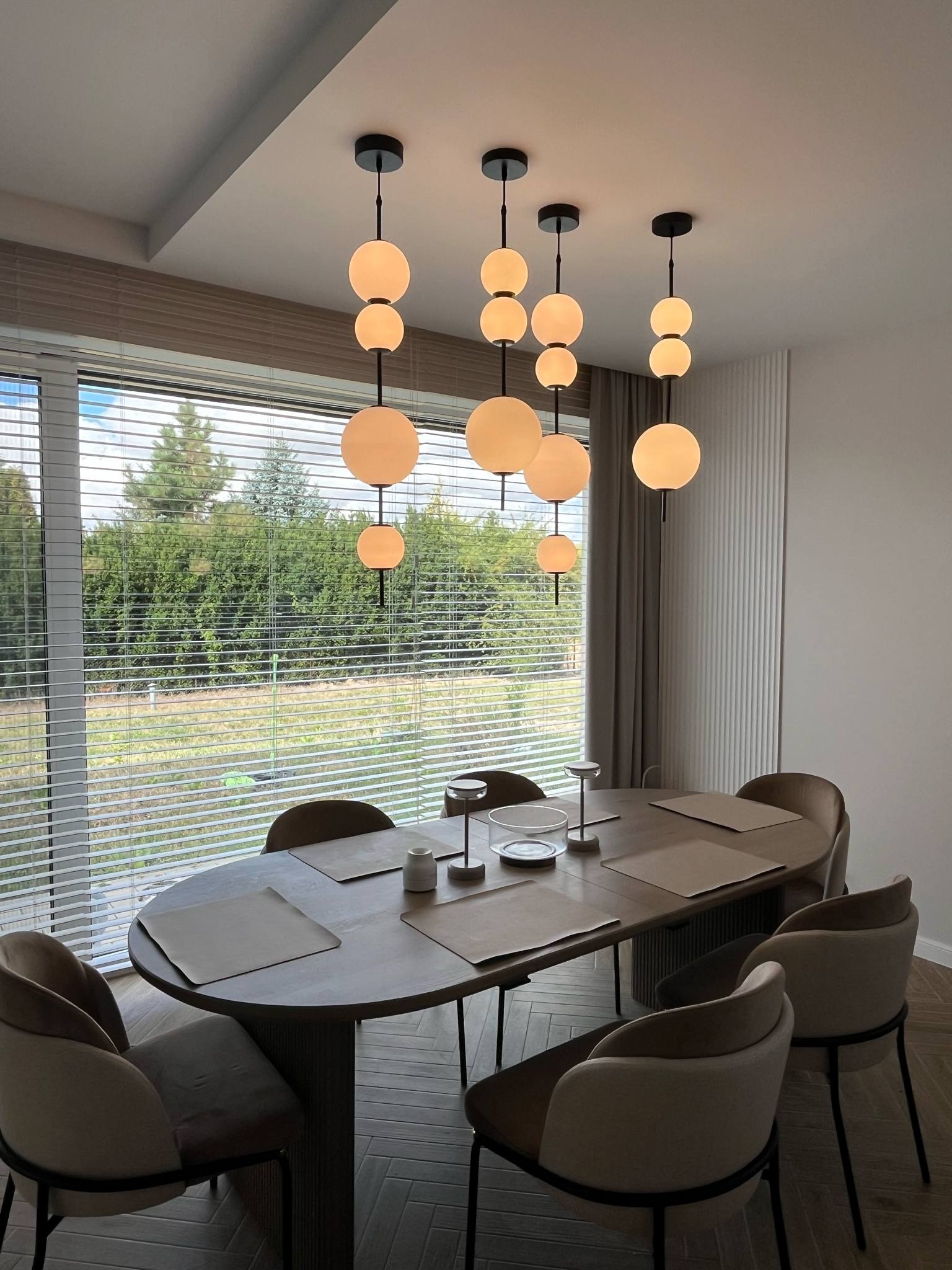 Nowoczesna jadalnia z owalnym stołem i designerskimi lampami kulami. Za oknem widok na zieleń, wnętrze utrzymane w stonowanej kolorystyce.