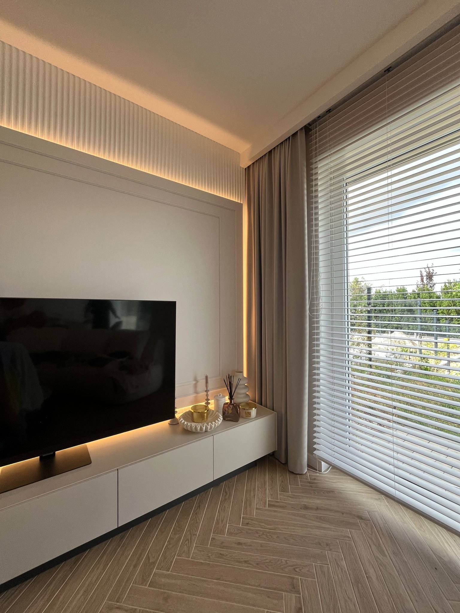Nowoczesny salon z telewizorem na szafce RTV, żaluzjami i parkietem w jodełkę. Ściana z dekoracyjną listwą i podświetleniem LED. Minimalistyczny design wnętrza.