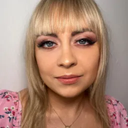 Portret kobiety z blond włosami i delikatnym różowym cieniowaniem powiek oraz wydłużonymi rzęsami, ubrana w kwiecistą bluzkę.