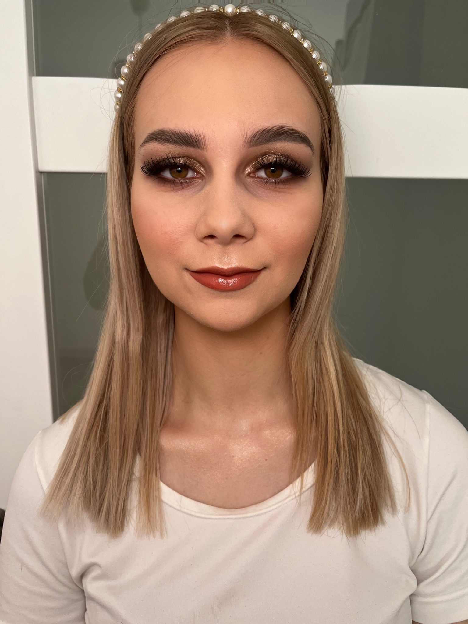 Portret kobiety z blond włosami, w białej koszulce, z pełnym, błyszczącym makijażem oczu w odcieniach złota i brązu, oraz błyszczącą pomadką w kolorze nude, z opaską z pereł na włosach.