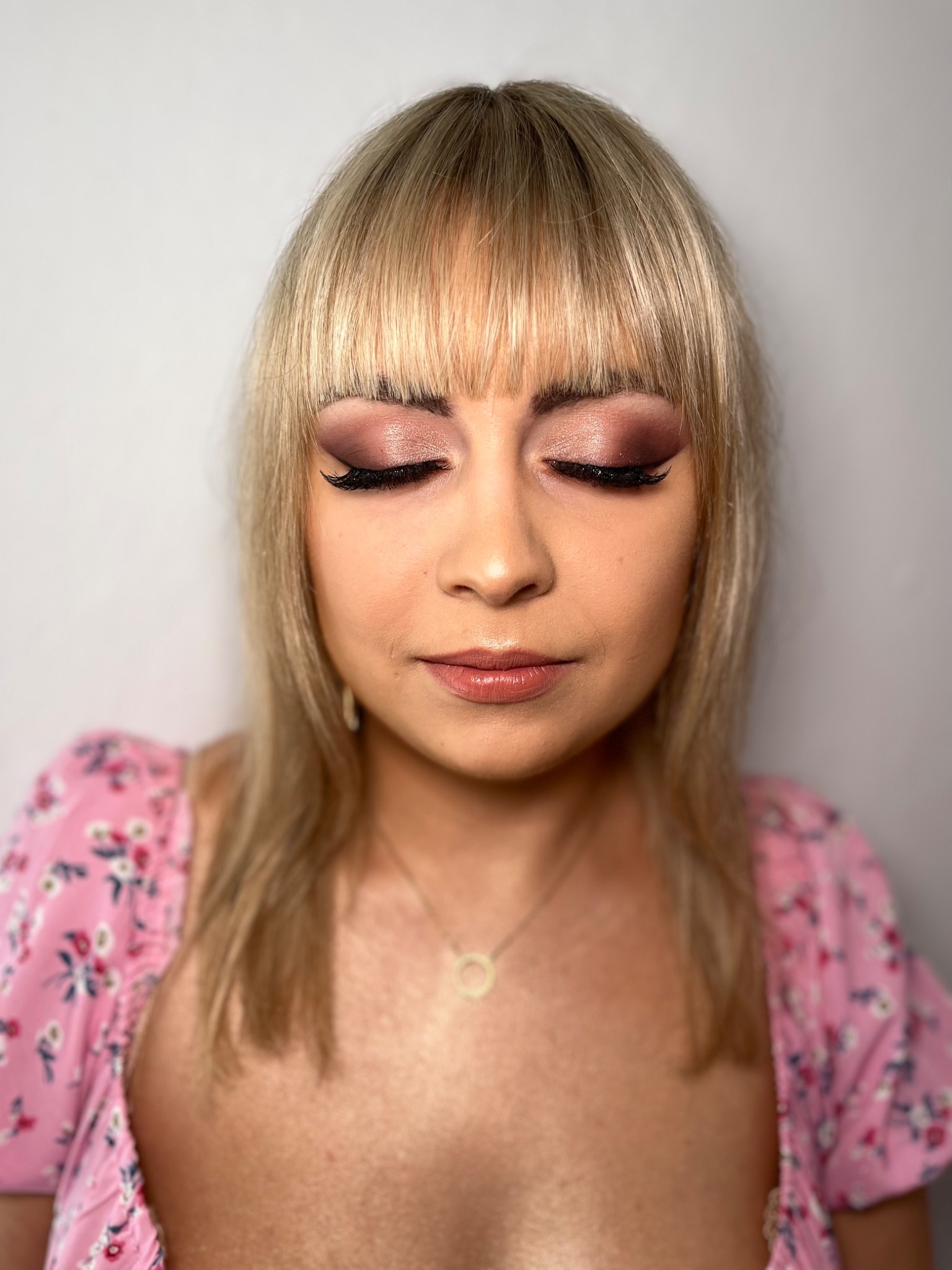 Portret kobiety z zamkniętymi oczami, prezentujący makijaż oczu w odcieniach różu i brązu z wyraźnie podkreślonymi rzęsami oraz delikatny makijaż ust w błyszczącym odcieniu nude, na tle jasnej ściany.