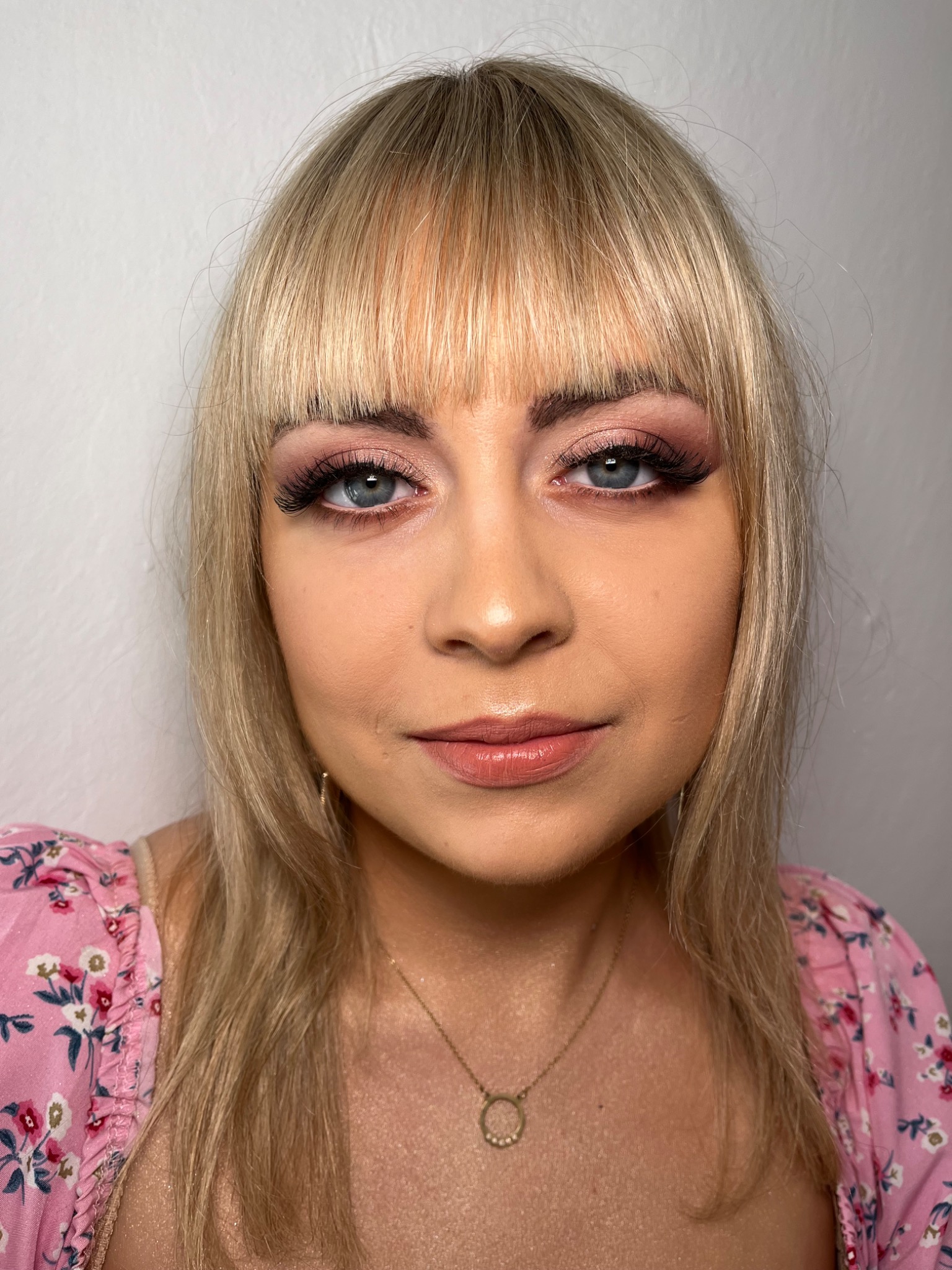 Portret kobiety z blond włosami i delikatnym różowym cieniowaniem powiek oraz wydłużonymi rzęsami, ubrana w kwiecistą bluzkę.