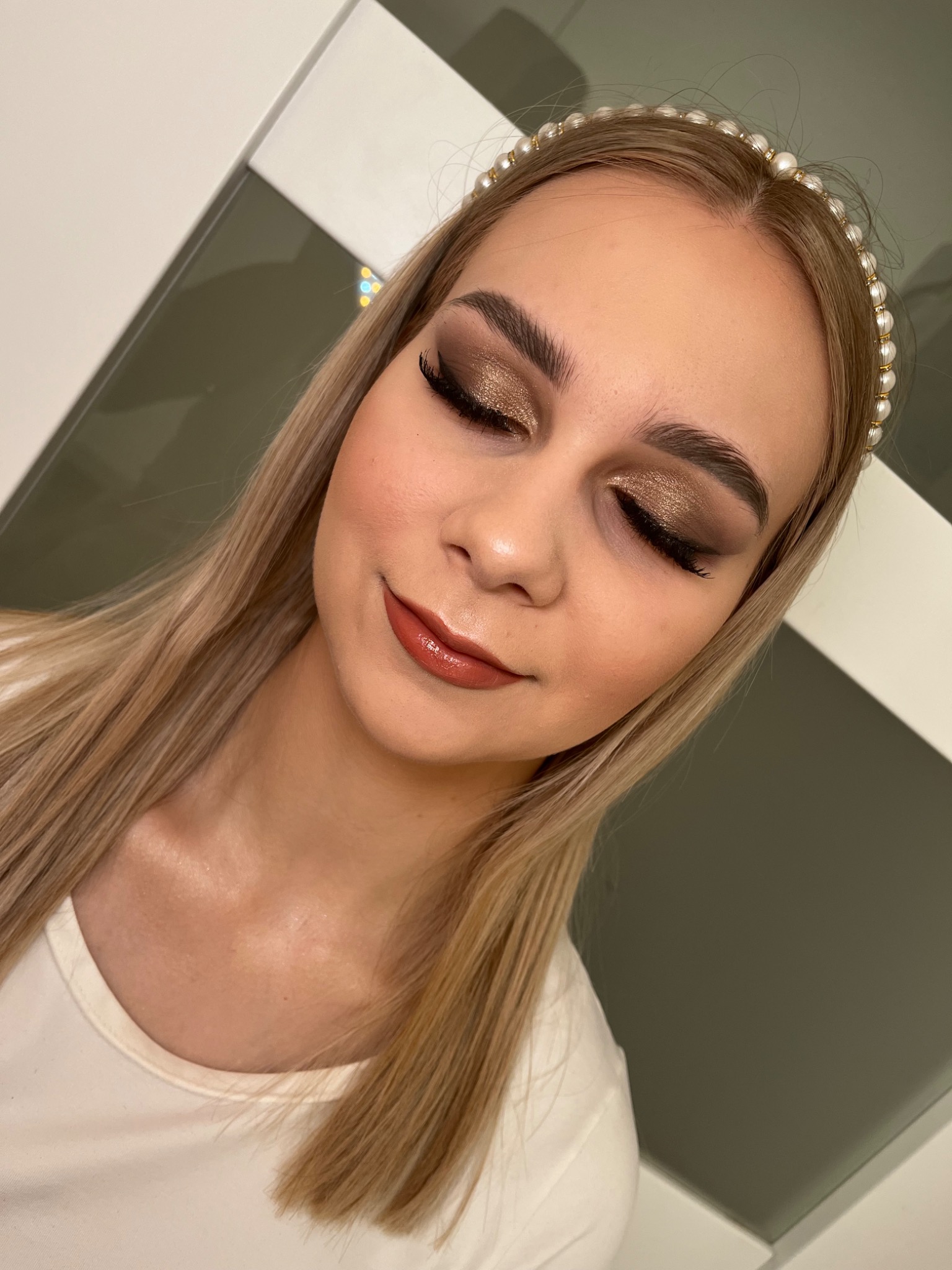 Portret kobiety z zamkniętymi oczami, prezentującej złoty, błyszczący makijaż oczu i brązową pomadkę, włosy blond ozdobione perłową opaską.