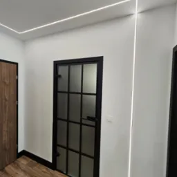 Nowoczesny korytarz z oświetleniem LED wzdłuż sufitu i ściany, czarne listwy przypodłogowe, drzwi z czarną ramą i szklanymi panelami, oraz drewniana podłoga.