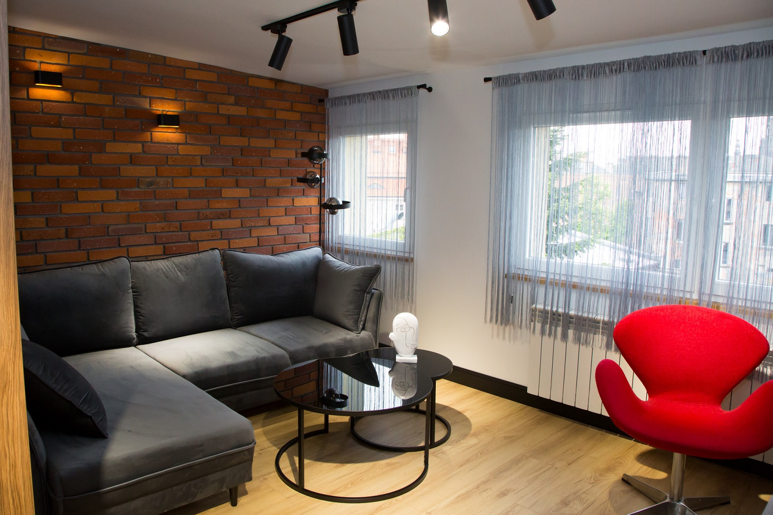 Salon z ceglaną ścianą, szarą sofą narożną, czarnym stolikiem kawowym, czerwonym fotelem i szarymi zasłonami sznurkowymi, oświetlony reflektorami sufitowymi i kinkietami.