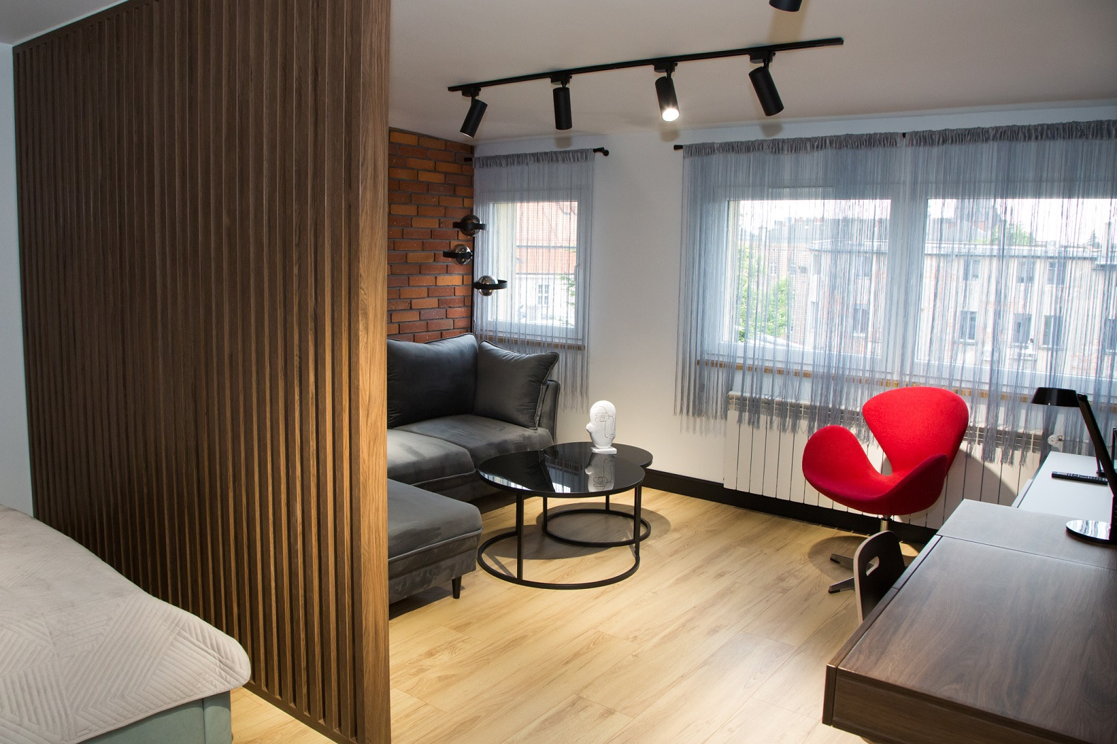 Nowoczesny salon z drewnianą ścianką działową, szarą sofą narożną, ceglaną ścianą, czarnym stolikiem kawowym i czerwonym fotelem Swan, oświetlony reflektorami sufitowymi.
