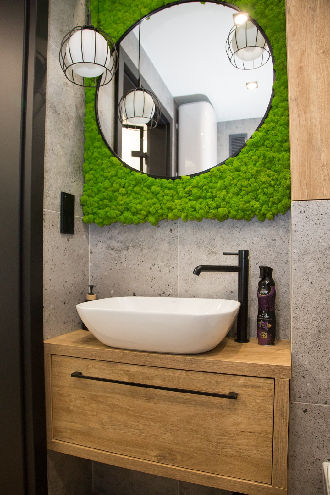 Nowoczesna łazienka z okrągłym lustrem otoczonym zielonym mchem, białą umywalką nablatową i czarną armaturą na szafce z drewnianym fornirem, wiszące lampy kuliste w tle odbicia.