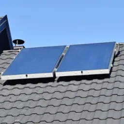 Dwa panele solarne z niebieskimi powierzchniami zamontowane na szarym dachu z dachówki ceramicznej pod błękitnym niebem.