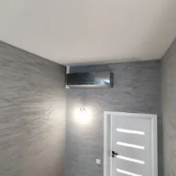 Nowoczesna, srebrna klimatyzacja ścienna w pomieszczeniu z dekoracyjną, szarą ścianą. Widoczna biała, frezowana ościeżnica drzwiowa z czarną klamką oraz kinkiet.