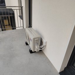 KARPATECH Dawid Szałda - Biała jednostka zewnętrzna klimatyzacji na balkonie, zamontowana na czarnych podkładkach antywibracyjnych, z widocznymi przewodami w białej osłonie.