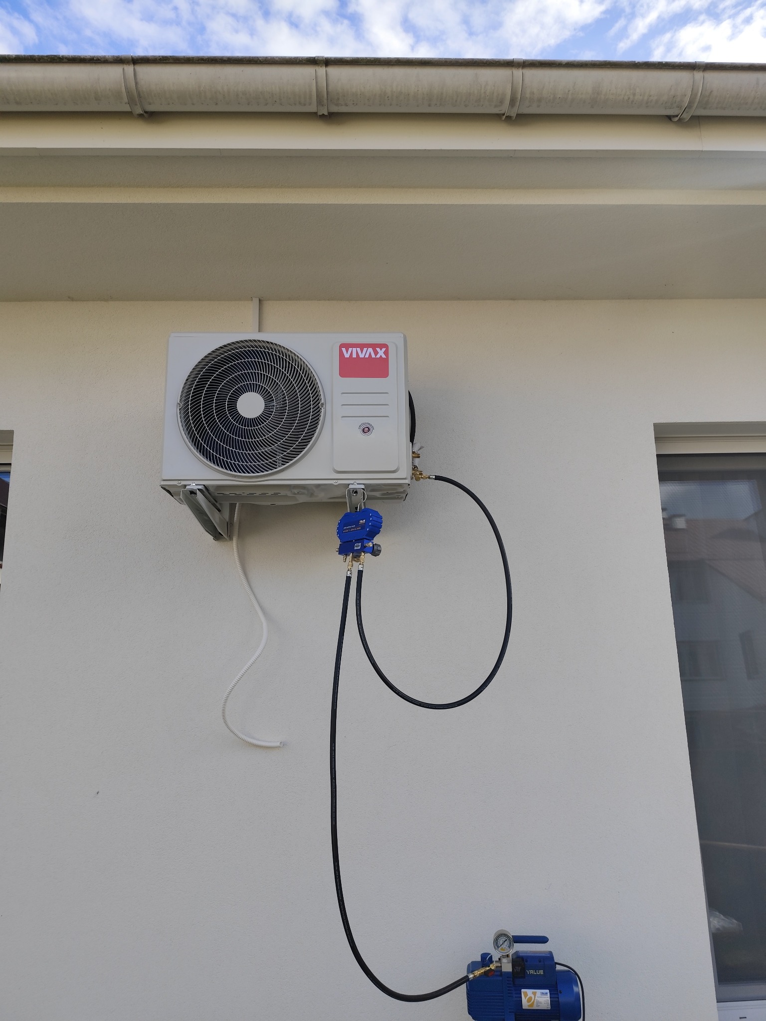 Zewnętrzna jednostka klimatyzacji marki Vivax zamontowana na białej ścianie budynku, podłączona do niebieskiej pompy próżniowej za pomocą czarnych przewodów.