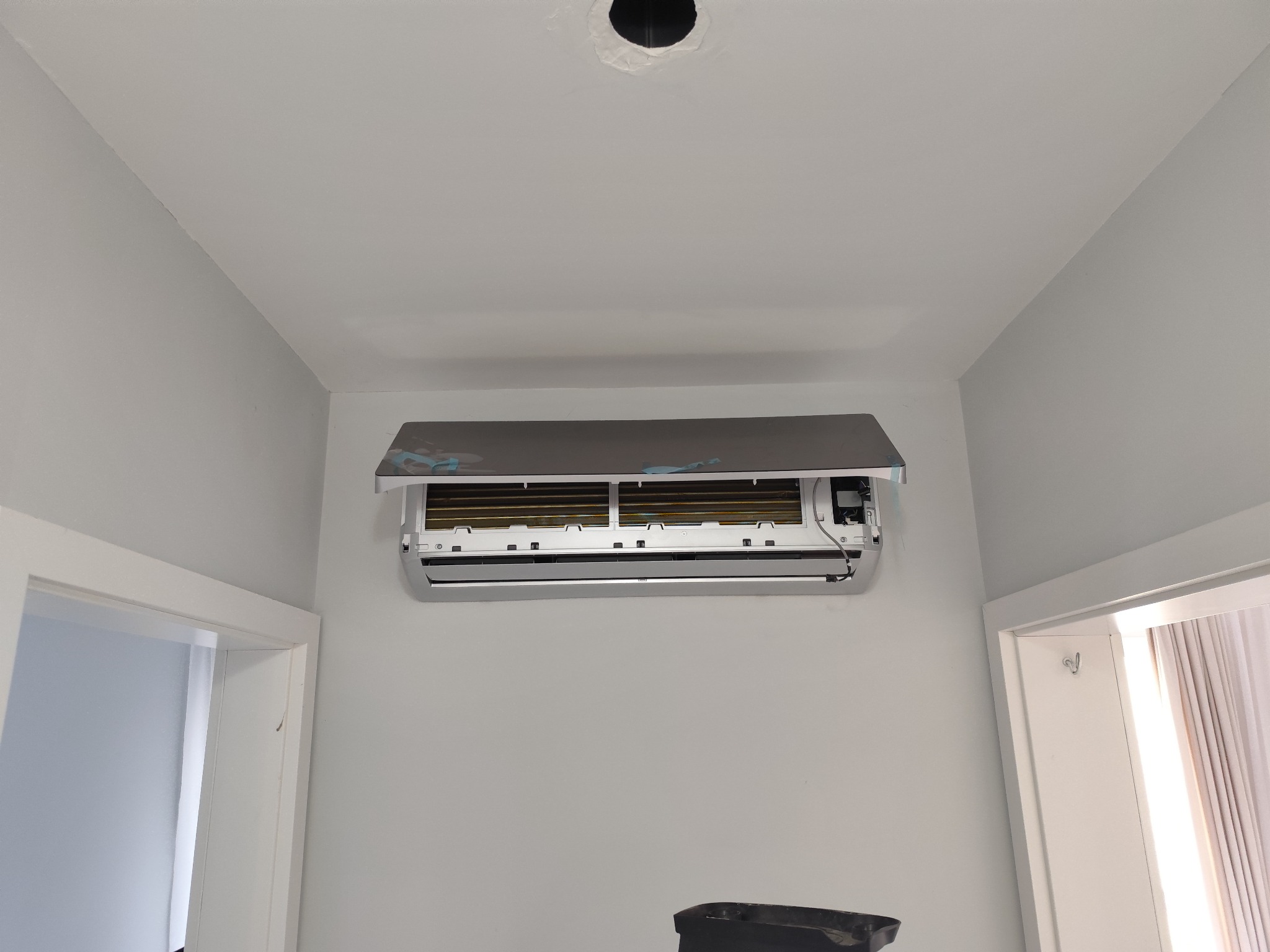 Klimatyzator ścienny w trakcie montażu, widoczne otwarte klapy i elementy instalacji, zarys otworu w suficie, zabezpieczenie folią ochronną.