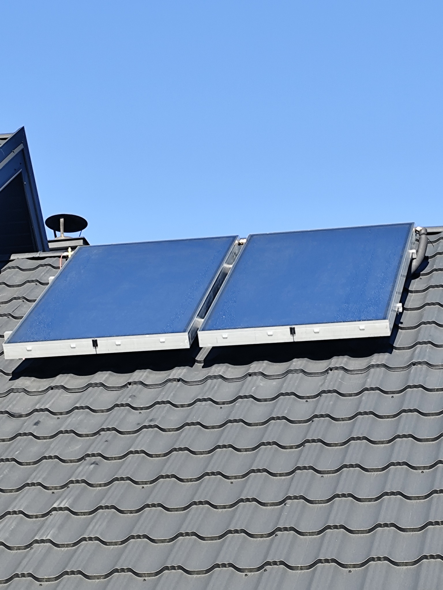 Dwa panele solarne z niebieskimi powierzchniami zamontowane na szarym dachu z dachówki ceramicznej pod błękitnym niebem.