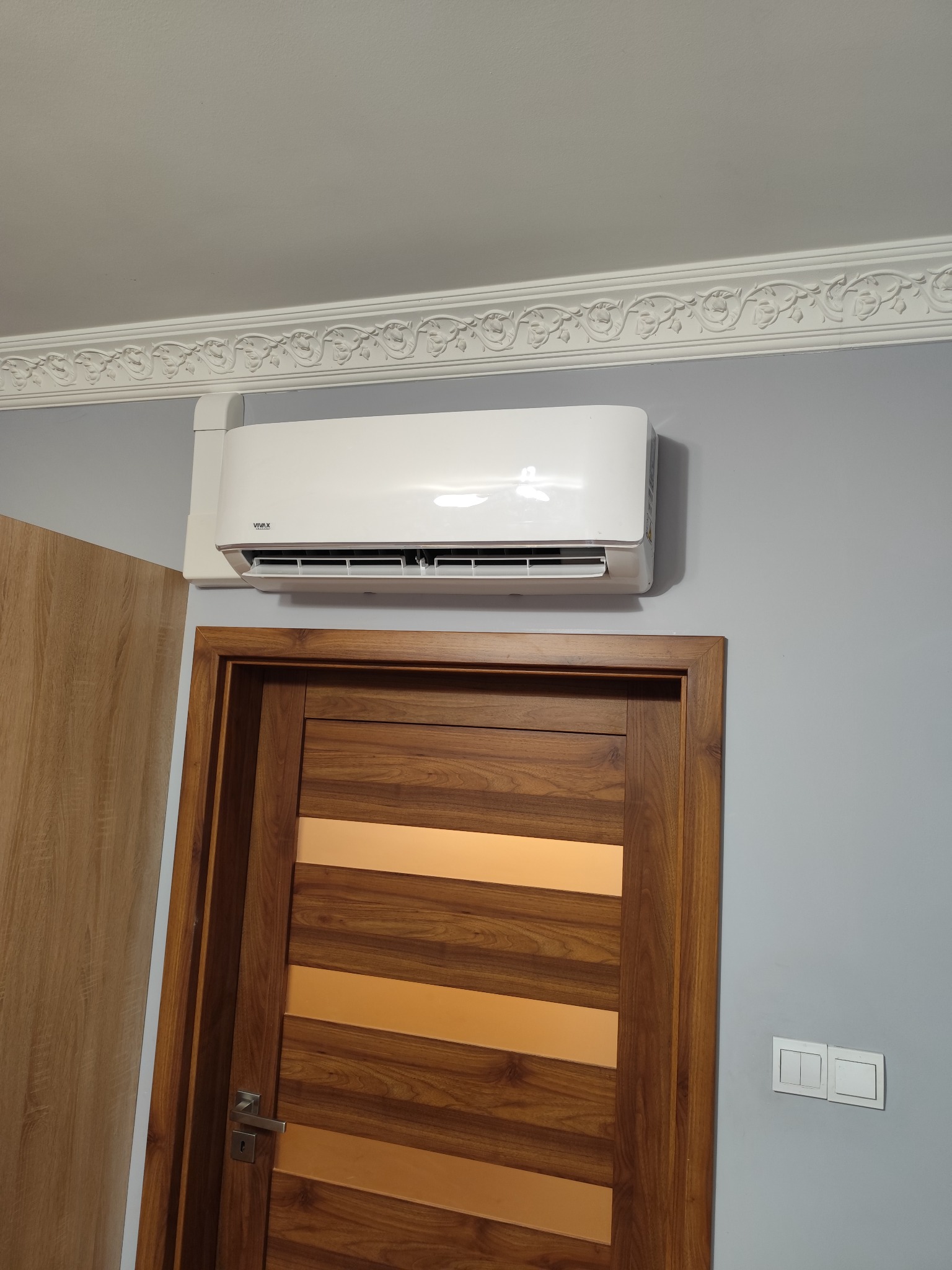 Biały klimatyzator VIVAX zamontowany nad drewnianymi drzwiami z poziomymi przeszkleniami, widoczne ozdobne listwy sufitowe i włącznik światła na szarej ścianie.