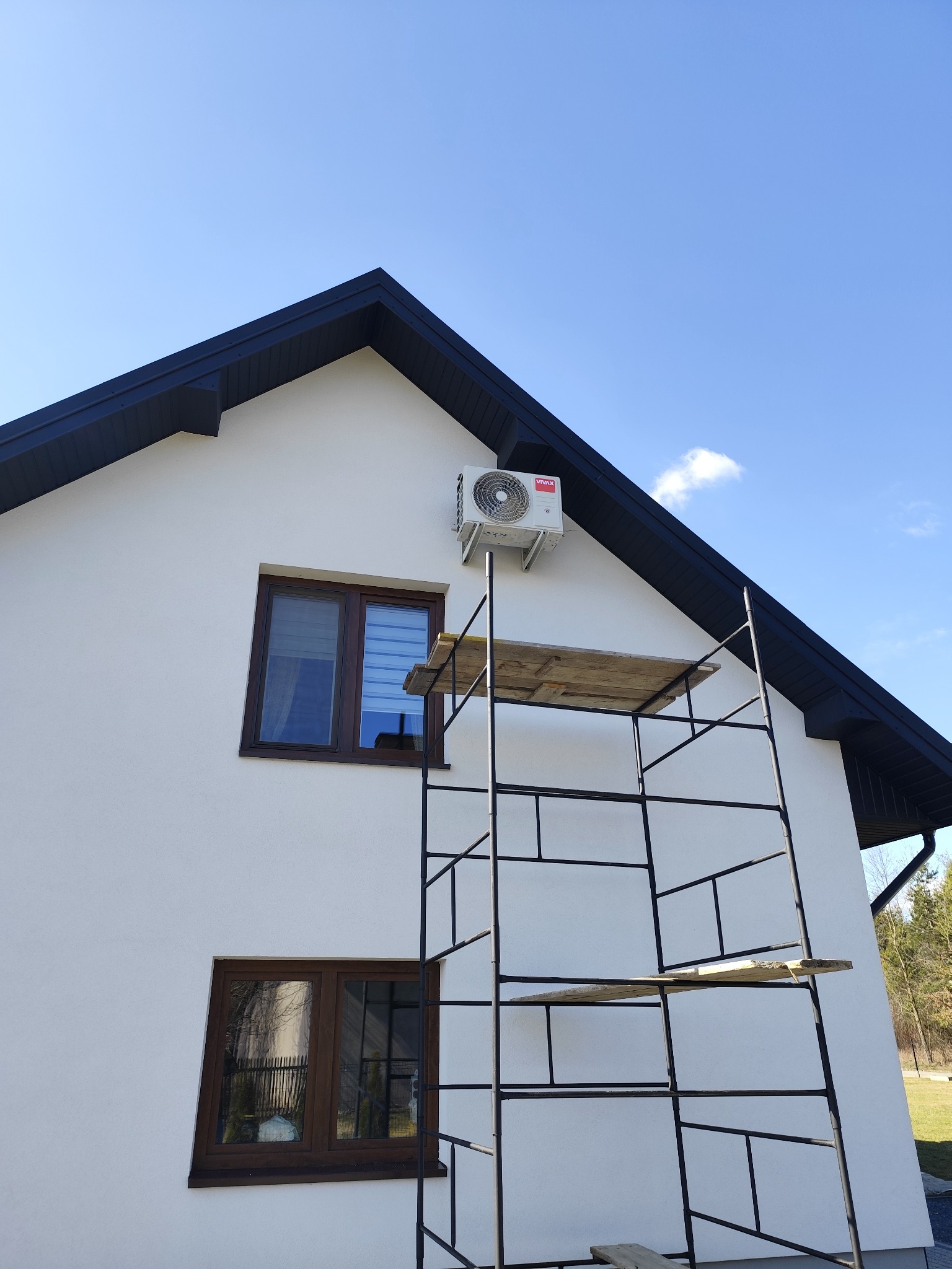 Biała elewacja domu z zamontowaną jednostką zewnętrzną klimatyzacji marki Vivax, widoczna z rusztowania ustawionego przy ścianie.