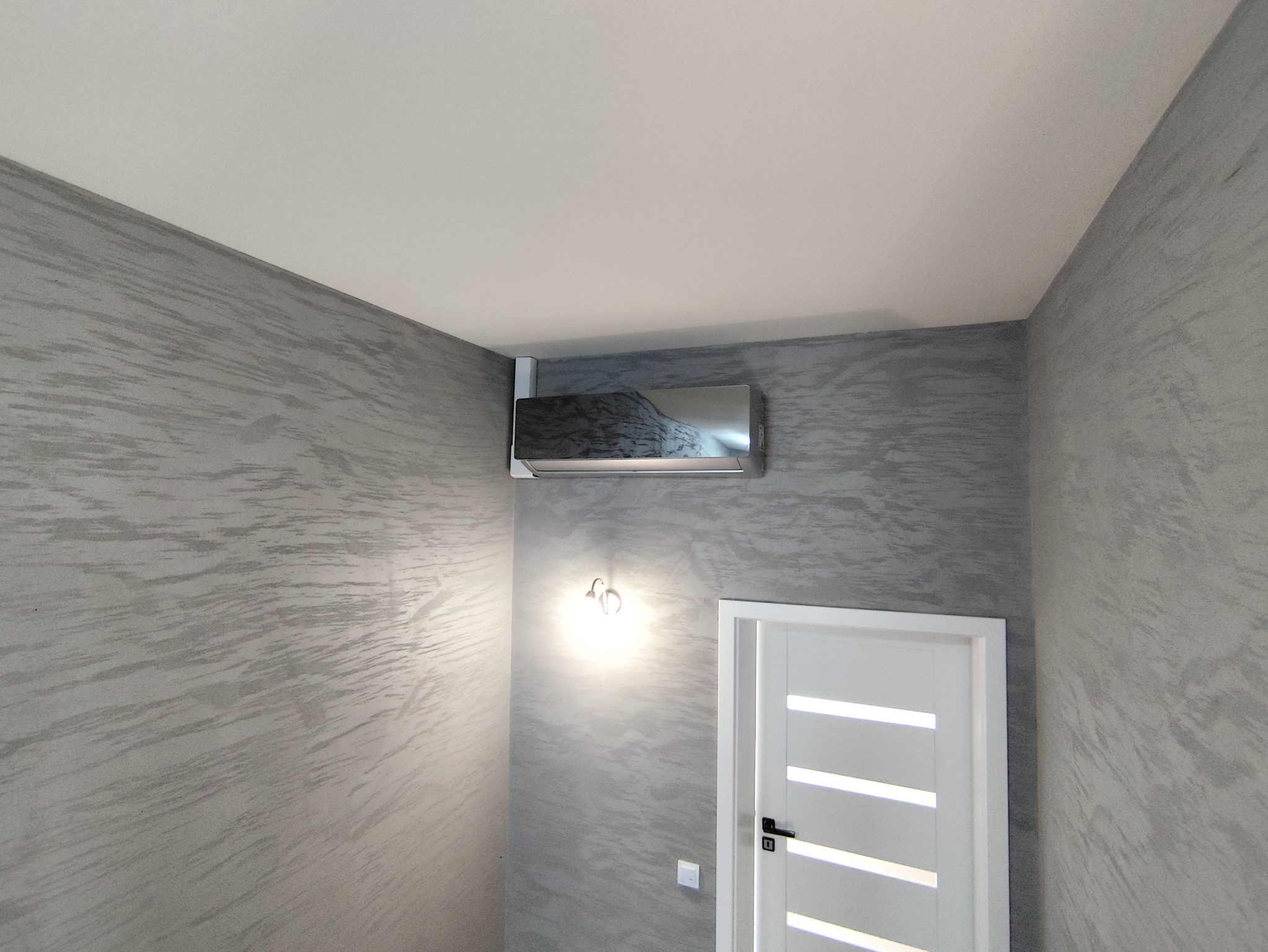 Nowoczesna, srebrna klimatyzacja ścienna w pomieszczeniu z dekoracyjną, szarą ścianą. Widoczna biała, frezowana ościeżnica drzwiowa z czarną klamką oraz kinkiet.