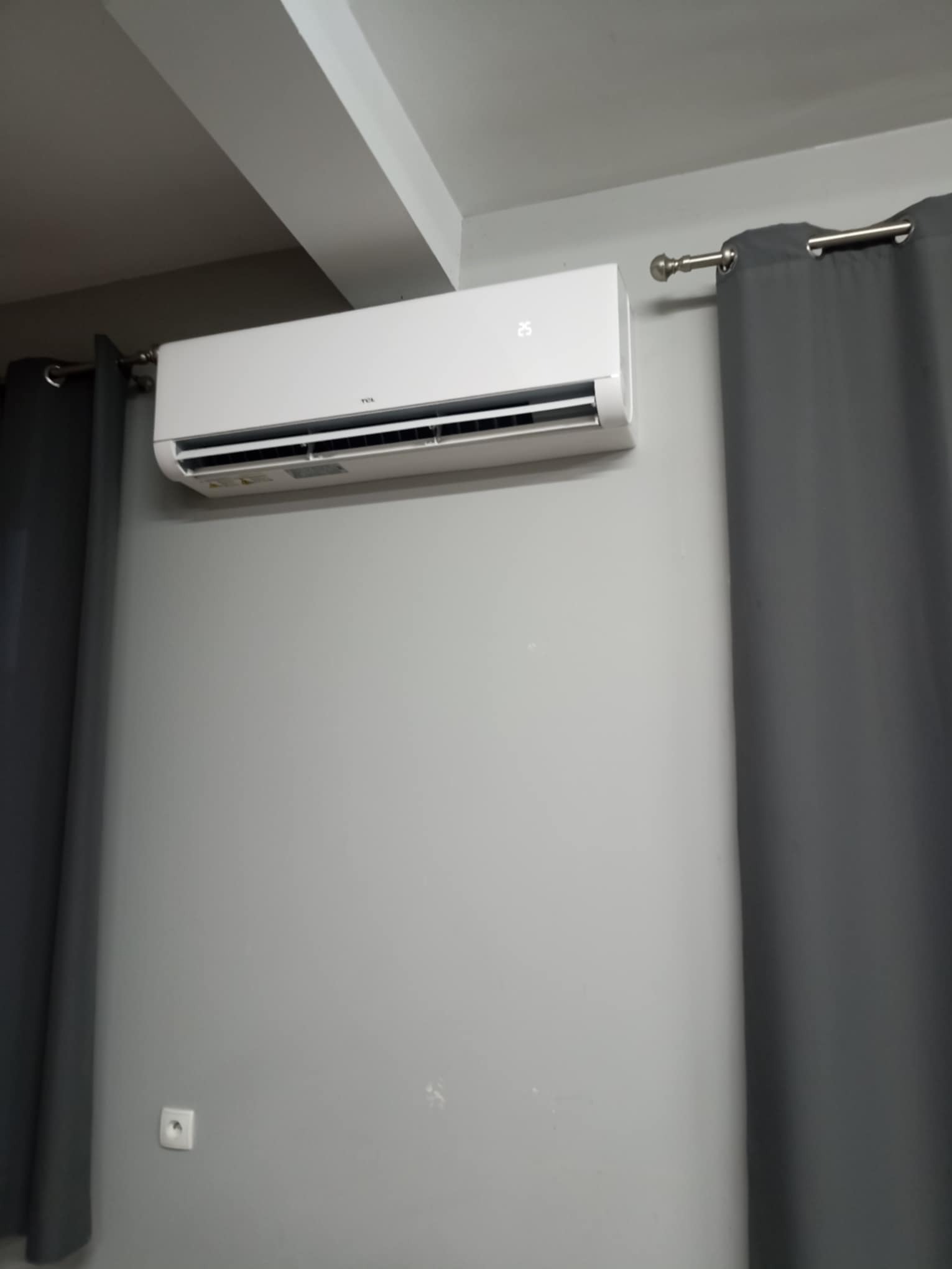 Biała klimatyzacja TCL zamontowana na szarej ścianie w pomieszczeniu z szarymi zasłonami. Widoczny fragment sufitu i gniazdko elektryczne.