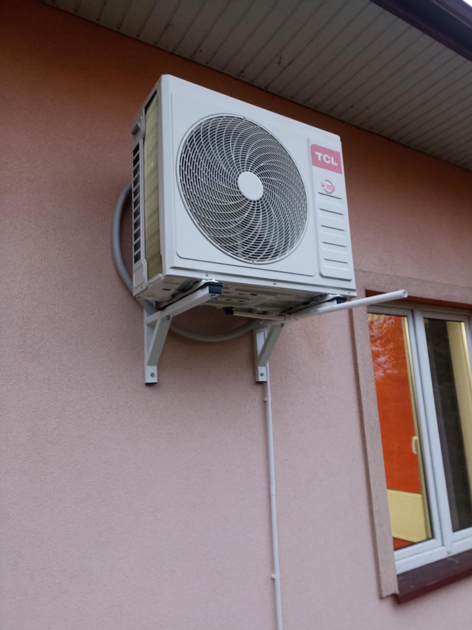 Biała jednostka zewnętrzna klimatyzacji TCL zamontowana na różowej ścianie budynku, widoczne rury i wsporniki. Okno z odbiciem w tle.