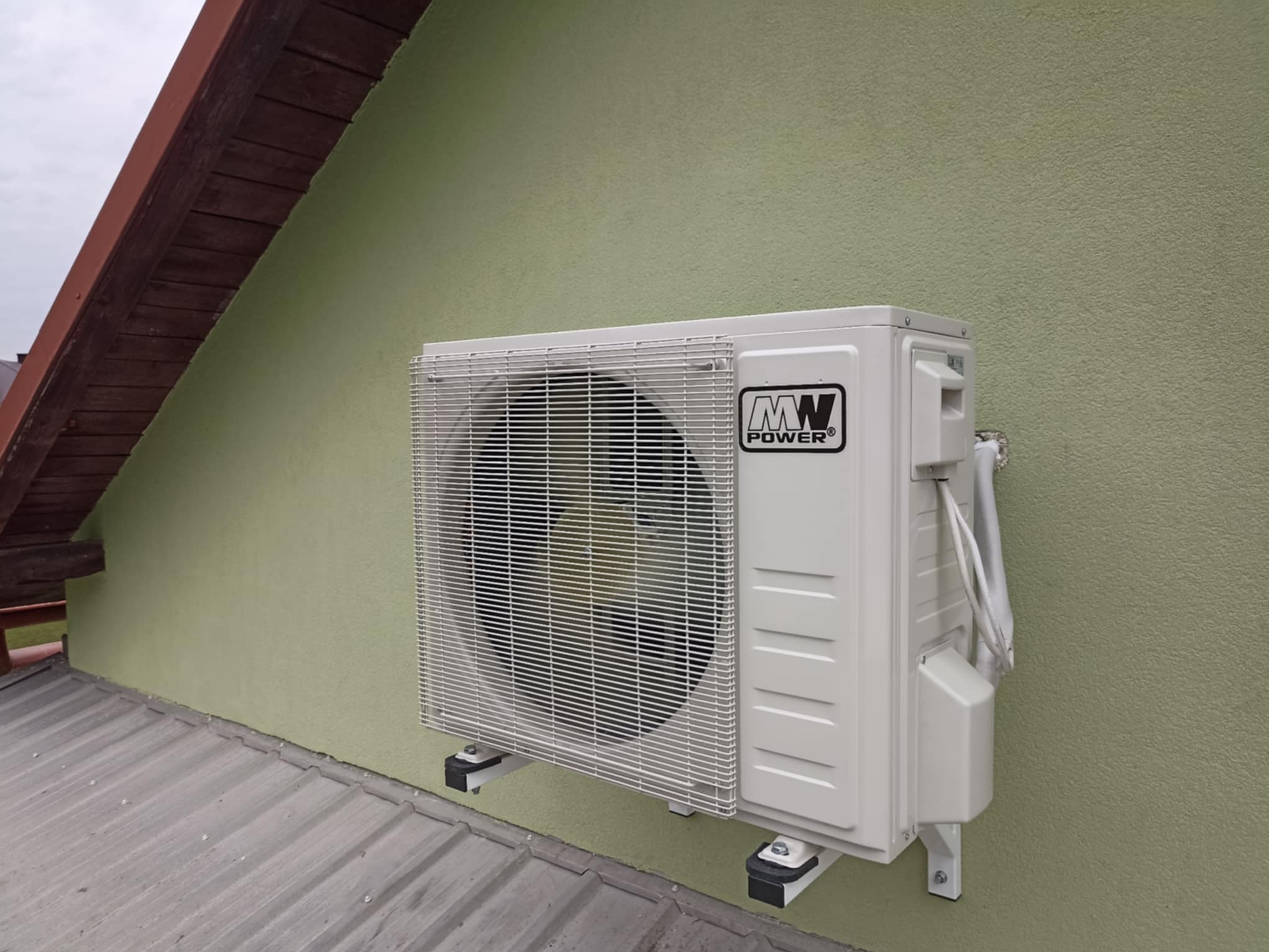 Biała jednostka zewnętrzna klimatyzacji MW Power zamontowana na zielonej ścianie budynku, widoczna z boku, z fragmentem dachu w tle.