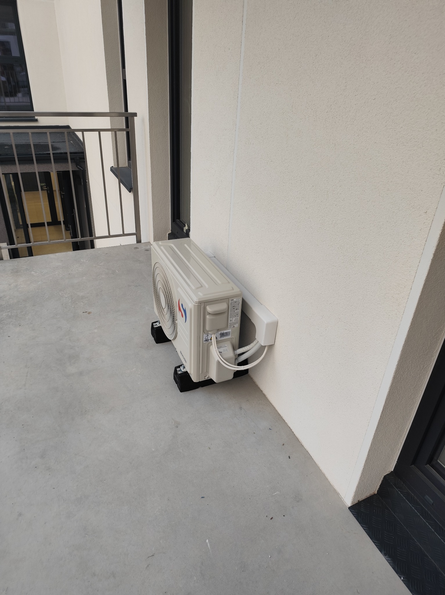 Biała jednostka zewnętrzna klimatyzacji na balkonie, zamontowana na czarnych podkładkach antywibracyjnych, z widocznymi przewodami w białej osłonie.