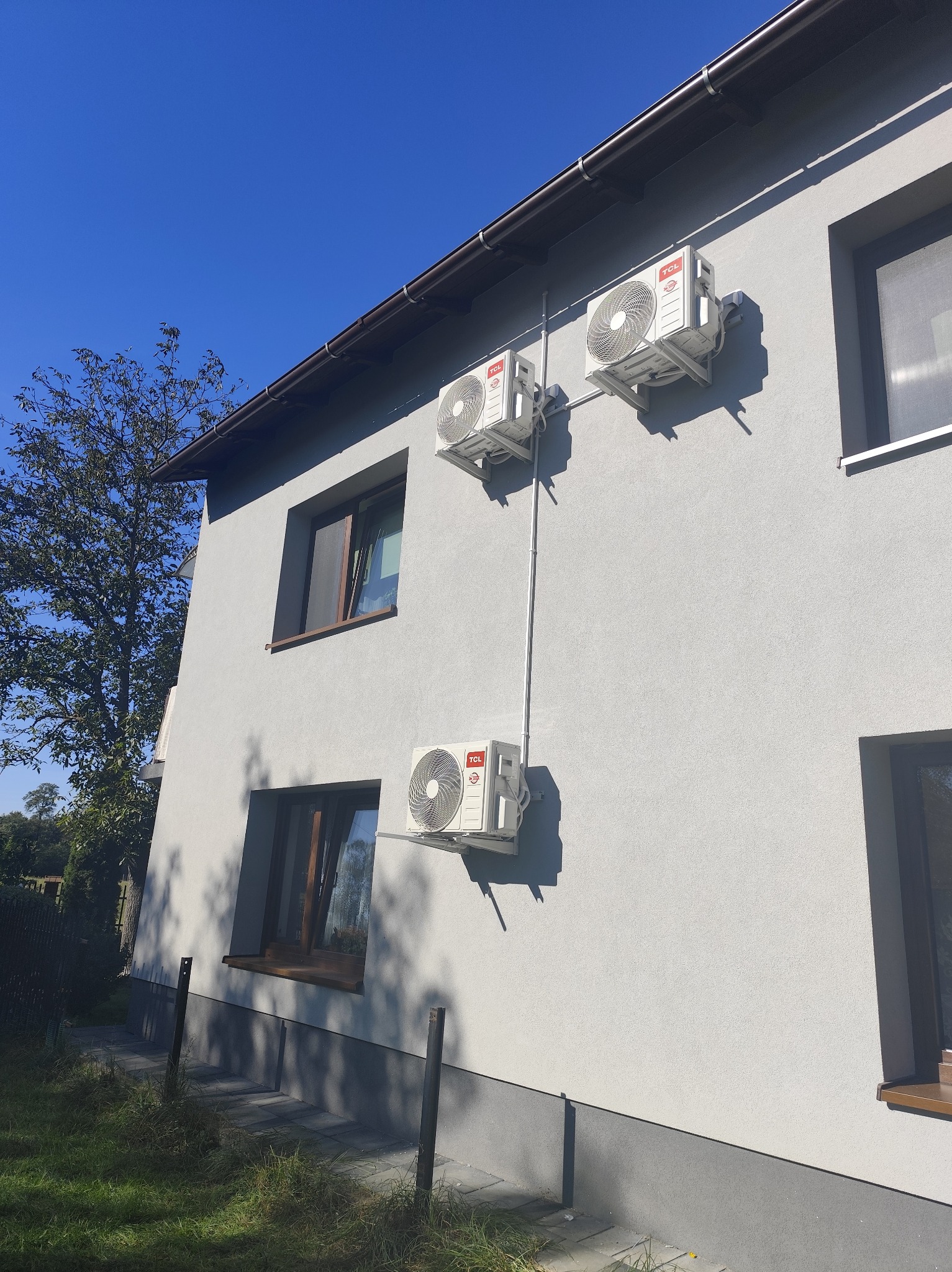 Elewacja domu z trzema zamontowanymi klimatyzatorami TCL, połączonymi rurami. Widok z dołu, błękitne niebo w tle. Instalacja klimatyzacji zewnętrznej.
