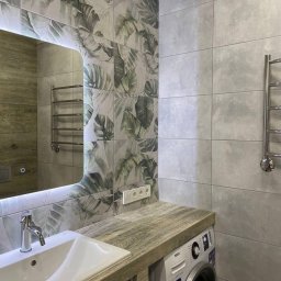 Blok.cz - Umywalka w łazience z płytkami w motywy roślinne, drewnopodobny blat i pralka pod blatem.