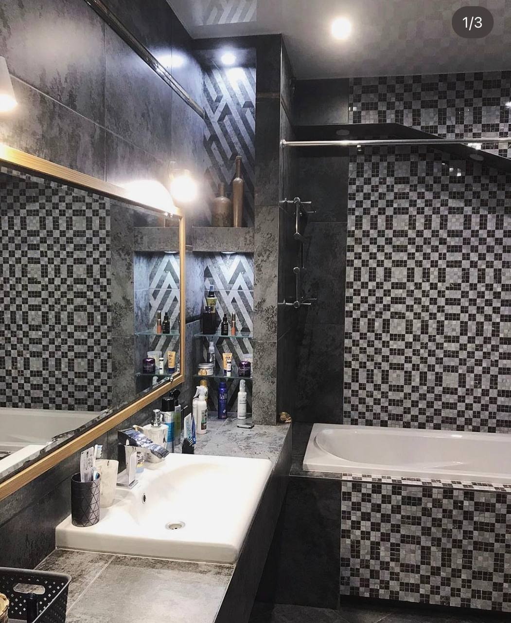 Nowoczesna łazienka z szarymi płytkami imitującymi kamień, mozaiką w kratkę, wnękami z półkami na kosmetyki i lustrem w złotej ramie.