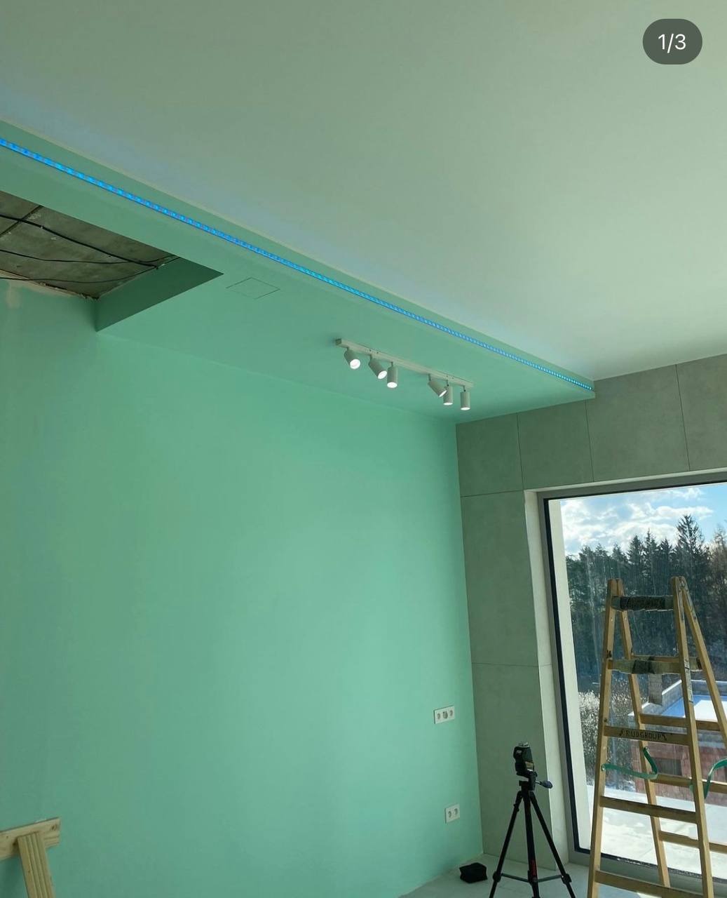 Wnętrze w trakcie remontu z pomalowanymi na zielono ścianami, widoczną listwą LED na suficie, lampami punktowymi, drabina przy oknie i kamera na statywie.