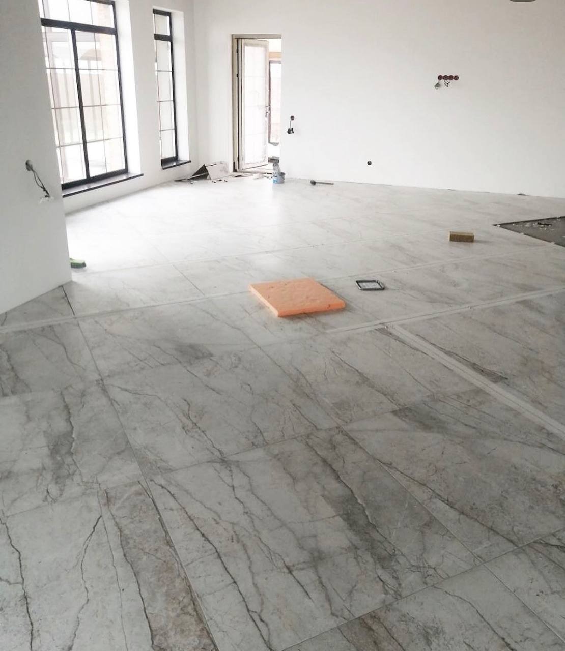 Układanie marmurowych płytek podłogowych w jasnym, przestronnym pomieszczeniu z wysokimi oknami; widoczna pomarańczowa podkładka i narzędzia glazurnicze w trakcie pracy.